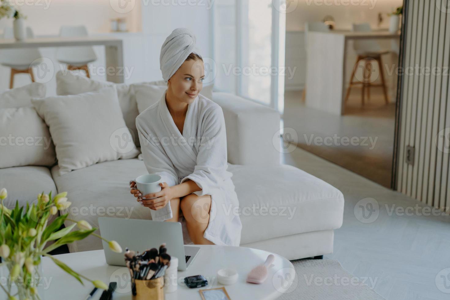 une femme réfléchie détendue vêtue d'un peignoir et d'une serviette enveloppée sur la tête est assise sur un canapé avec une tasse de boisson près de la table avec des produits cosmétiques a l'air pensif à côté pose contre l'intérieur confortable de la maison photo