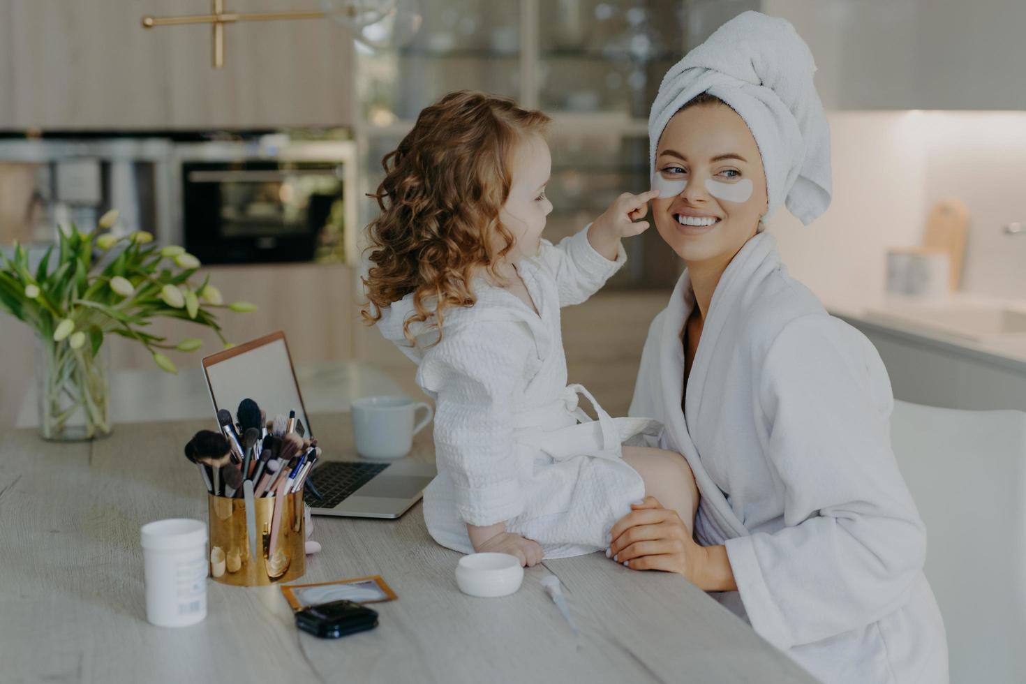 jolie petite fille aux cheveux bouclés regarde comment la mère fait les procédures cosmétiques applique des patchs de collagène sous les yeux pour le traitement de la peau porte des peignoirs pose près du bureau avec des produits de beauté. soins quotidiens photo