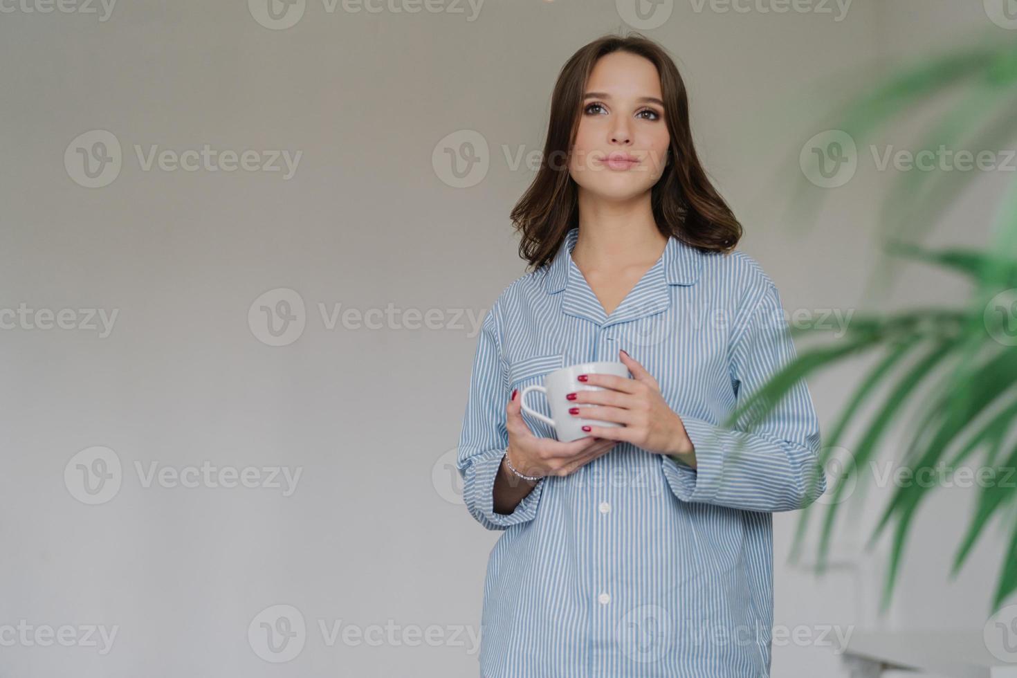 photo horizontale d'une jolie femme vêtue d'une merde rayée surdimensionnée, a des toilettes attrayantes, tient une tasse de café ou de latte, se tient à l'intérieur avec un espace de copie vierge à gauche pour votre contenu ou texte publicitaire