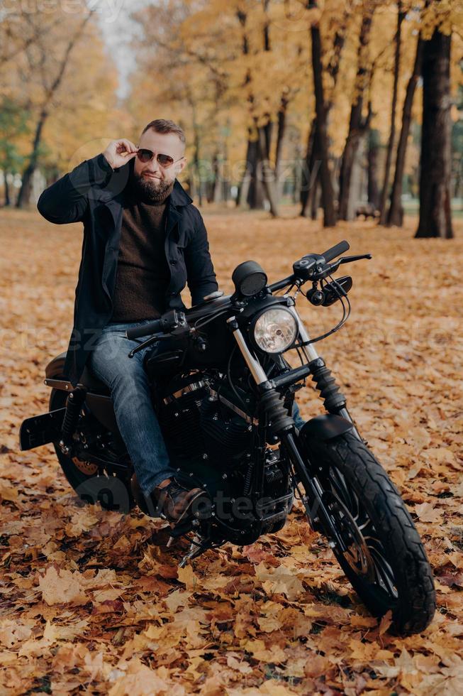 image verticale d'un motard réfléchi porte des lunettes de soleil, fait de la moto dans le parc, pose dans la rue, porte des vêtements élégants, aime le temps libre et les loisirs, admire la nature, profite du temps automnal ensoleillé photo