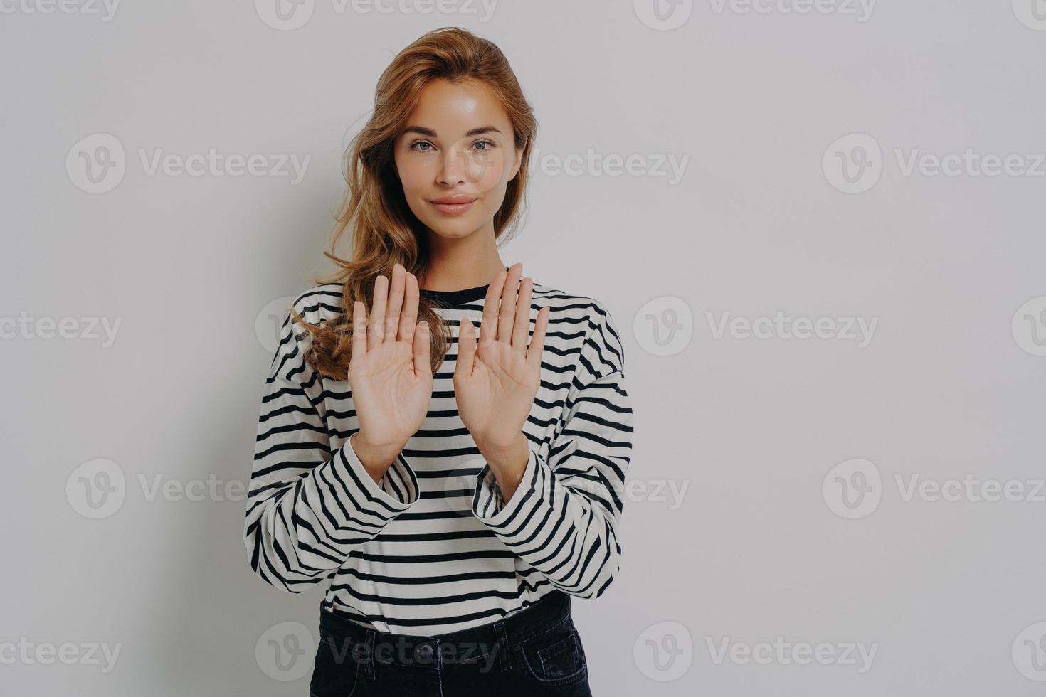 jeune femme confiante gesticulant panneau d'arrêt, disant non tout en se tenant isolé contre le mur gris photo