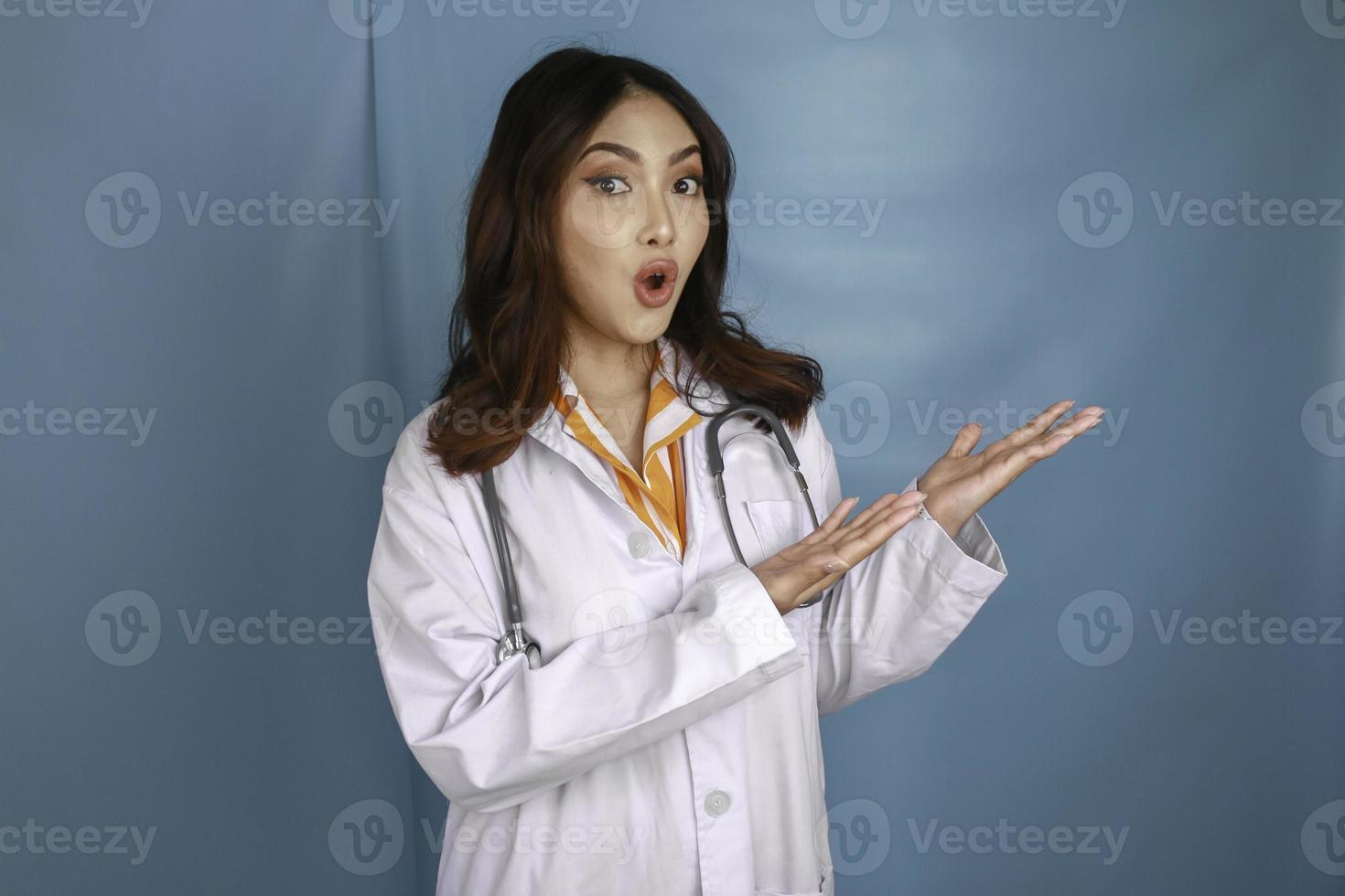 portrait d'une jeune femme médecin asiatique, professionnel de la santé est choqué et pointe vers un espace de copie isolé sur fond bleu photo