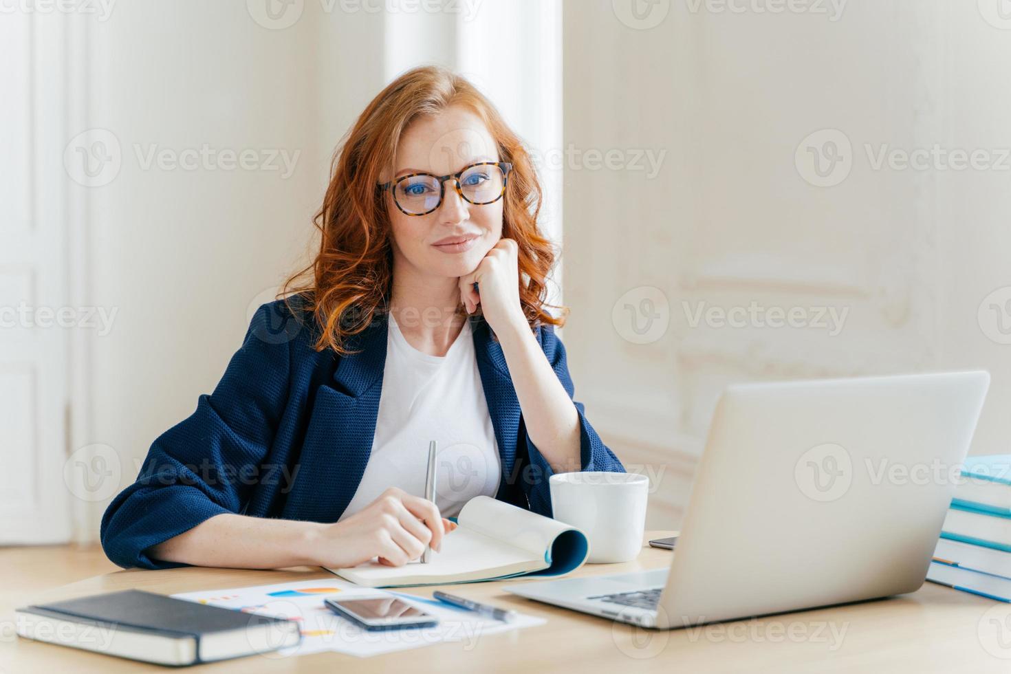 une employée de bureau rousse bouclée analyse les données, établit un rapport comptable, pose dans un bureau de coworking, améliore les ventes en ligne, boit du café, porte des lunettes et des vêtements de cérémonie. employée à l'intérieur photo