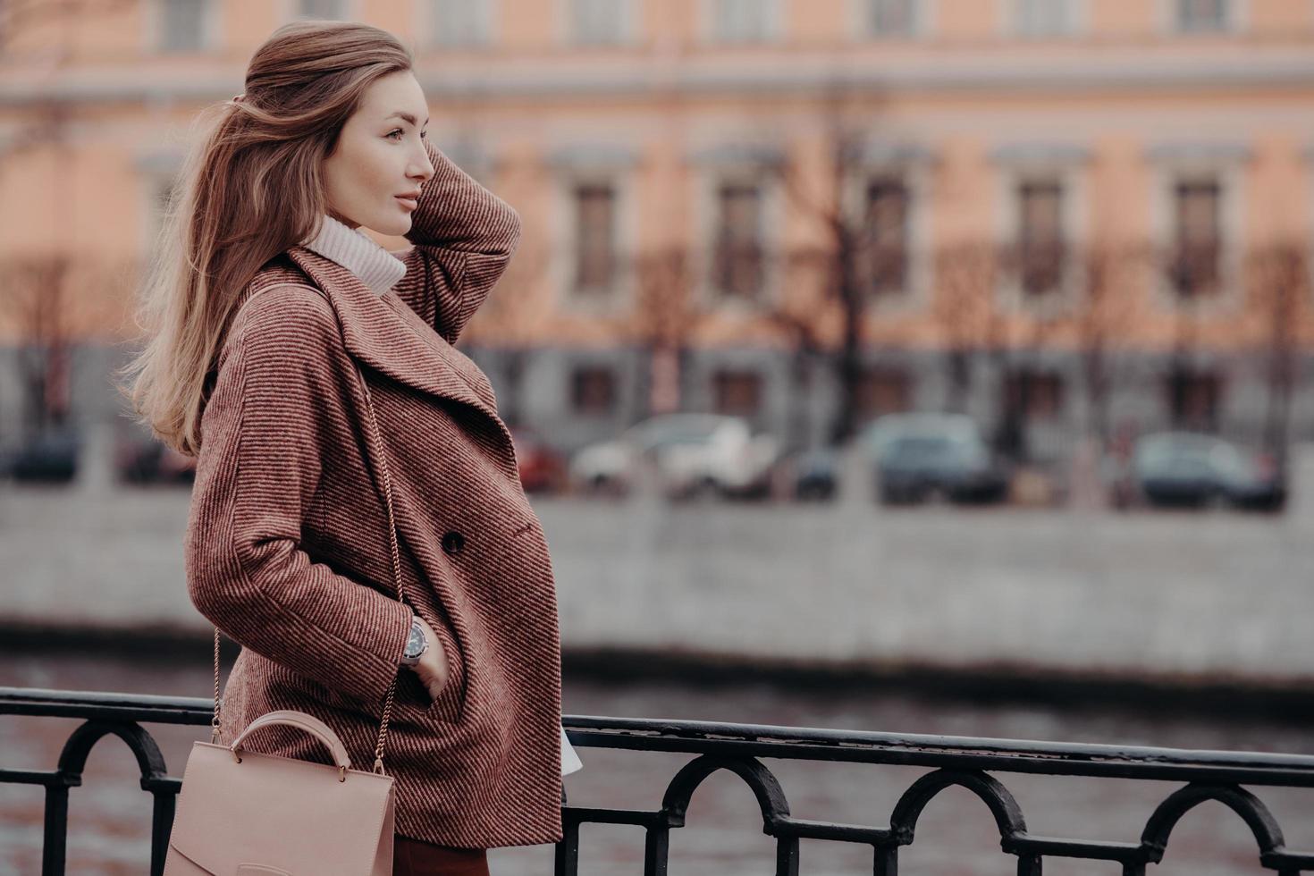 manteau à la mode, belle femme vêtue d'un manteau élégant, garde la main dans la poche, se concentre sur la distance, se promène en ville, pense à quelque chose, se repose bien. concept de personnes et de style de vie. photo