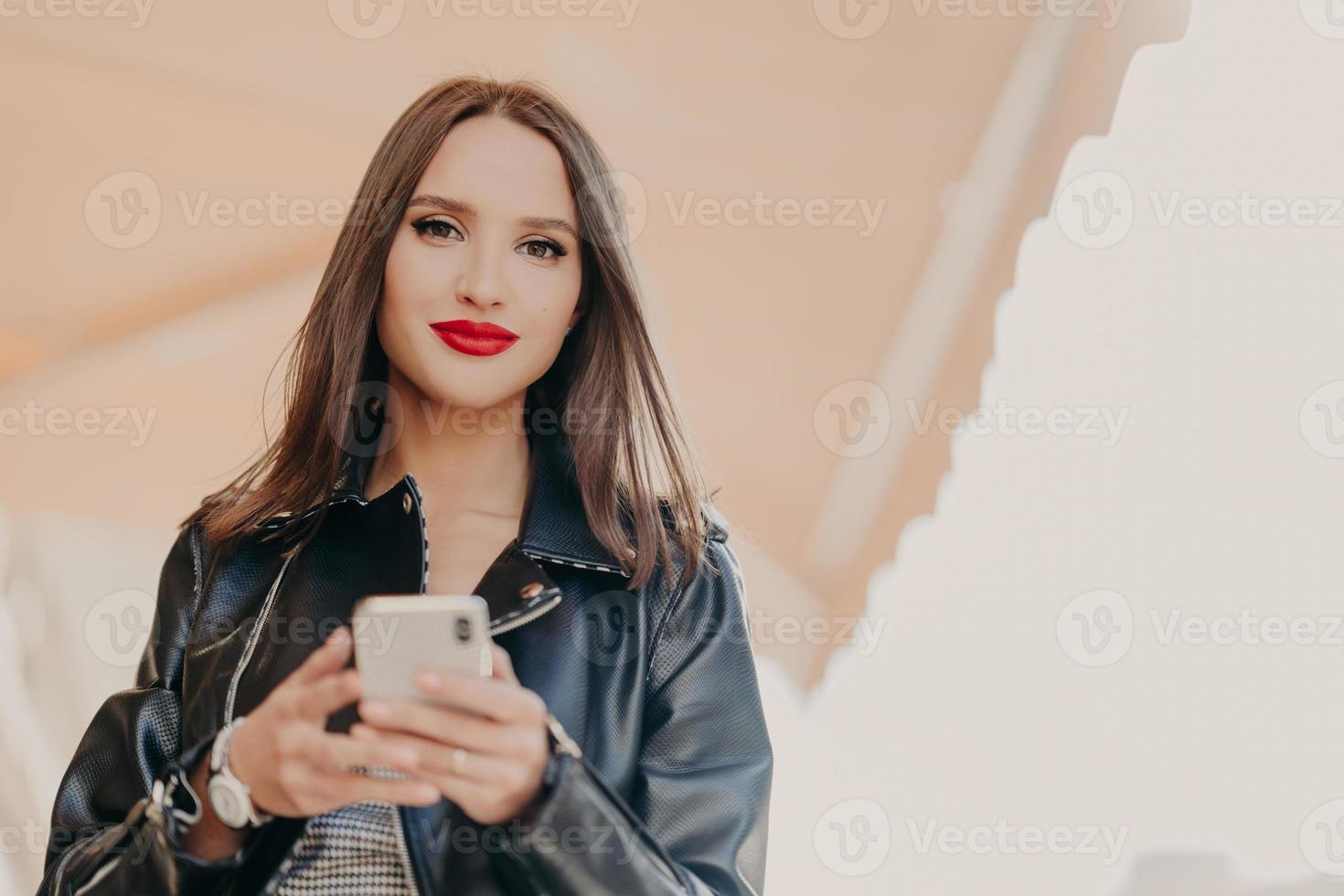 photo horizontale d'une jolie femme aux cheveux noirs avec des lèvres peintes en rouge, vêtue d'une veste en cuir noir, tient un téléphone portable moderne, envoie des messages texte, discute en ligne, se promène dans la rue de la ville. La technologie