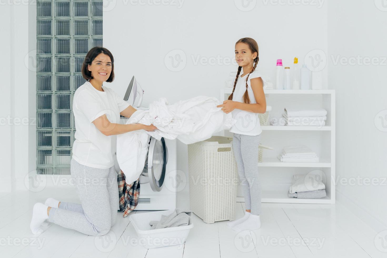 heureuse femme au foyer fait la lessive avec une petite aide adorable. la mère et la fille lavent les vêtements dans la buanderie, chargent le linge dans la laveuse. la femme se tient à genoux près de la machine à laver. notion de ménage photo