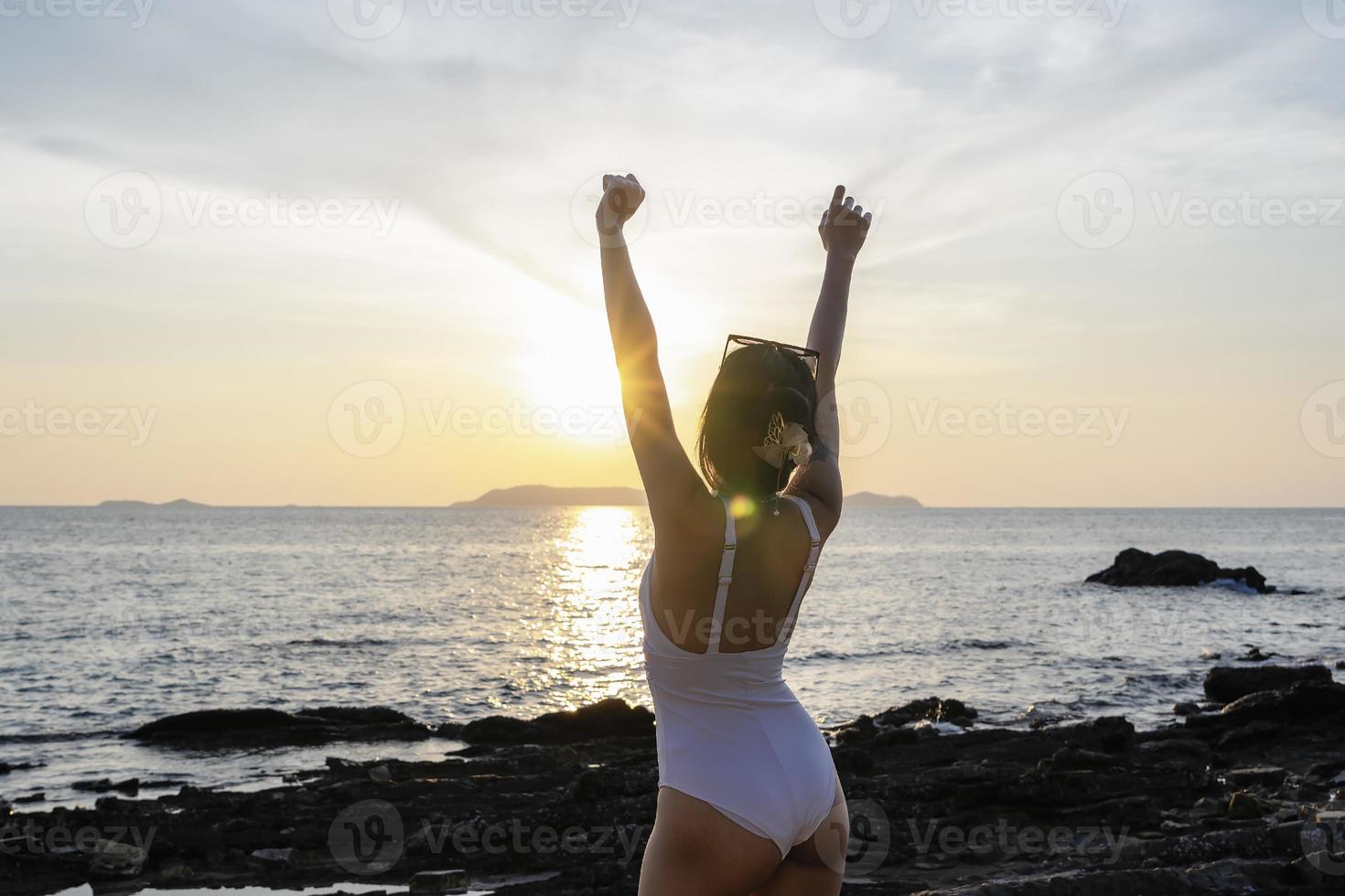 femme touriste levant les mains avec coucher de soleil sur la plage en voyage de vacances. femme portant un maillot de bain relaxant et bras levés avec des grains de sable sur la peau. dame asiatique profitant de la nature en vacances. notion de liberté photo