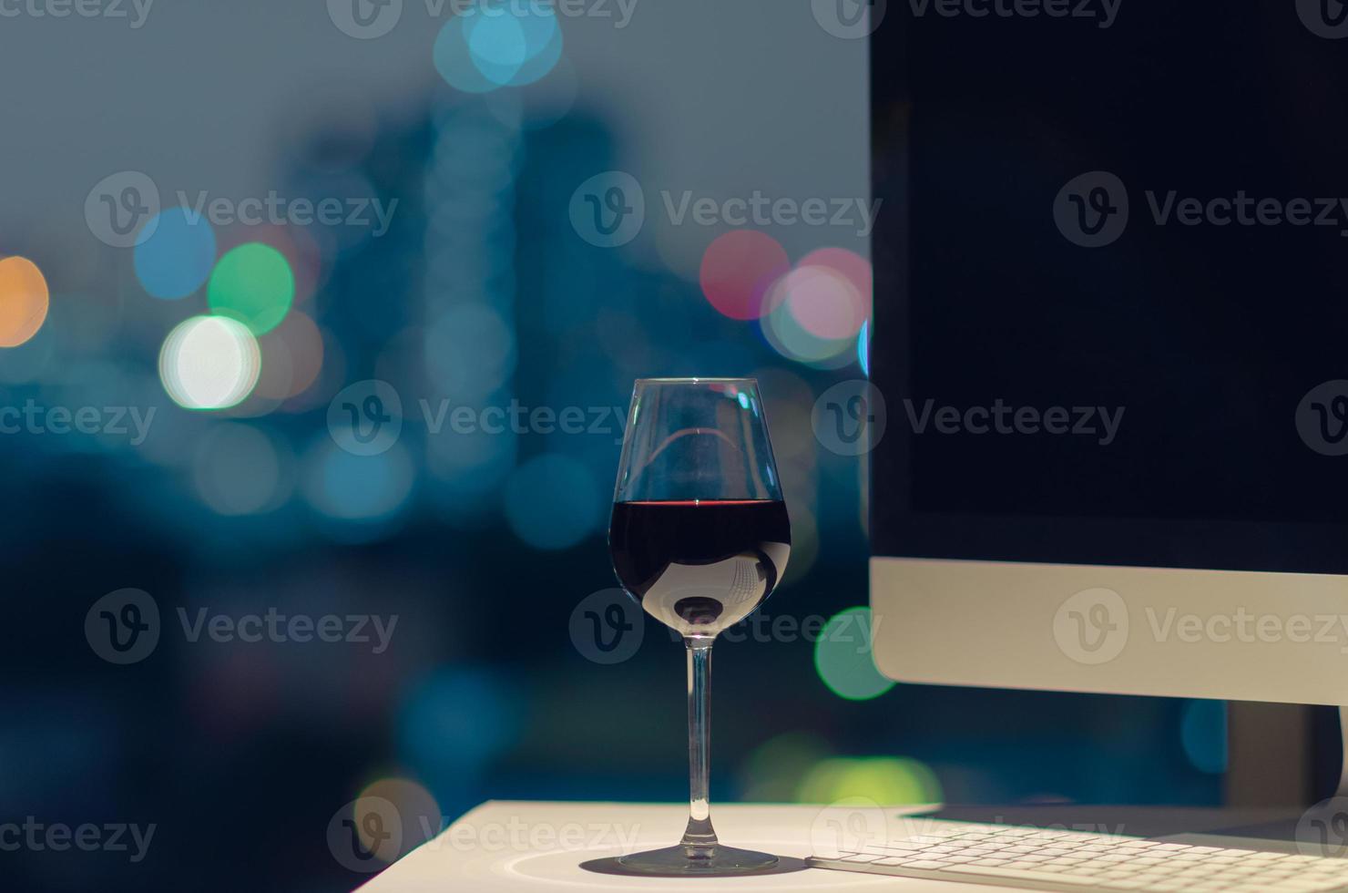 un verre de vin rouge posé sur une table de travail, un écran d'ordinateur et un clavier avec bokeh de lumières de la ville. photo