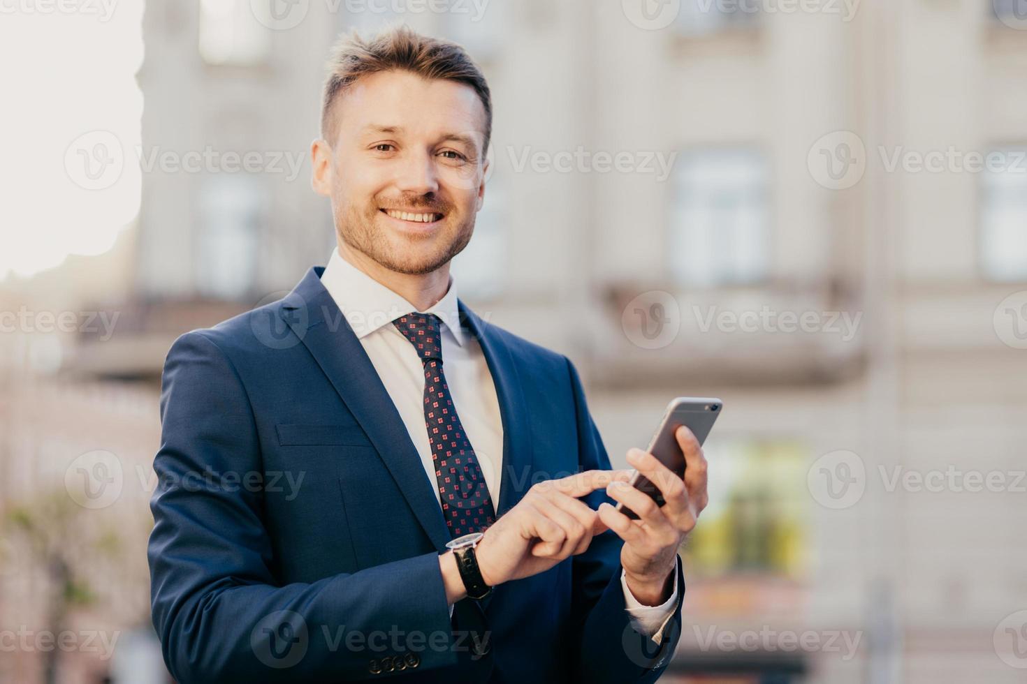 un homme d'affaires satisfait tient un smartphone, lit les e-mails de l'investisseur, heureux de recevoir de bonnes nouvelles sur l'entreprise, se tient en territoire urbain, porte un costume noir et une cravate élégante. notion de technologie photo