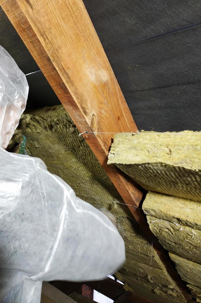 un homme dans une combinaison de protection met de la laine minérale entre la poutre du toit de la maison pour son réchauffement du froid photo