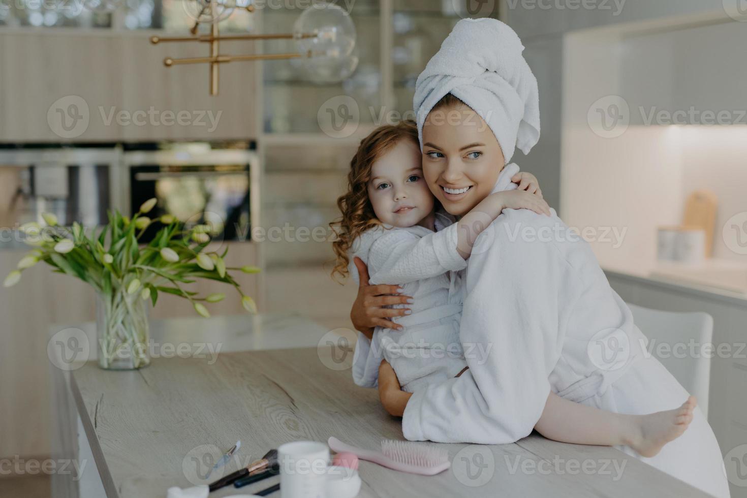 photo de la jeune mère et de sa fille s'embrassent et expriment leur amour l'une à l'autre posent dans un appartement moderne portent des peignoirs de bain blancs doux vont avoir des procédures cosmétiques ont une peau saine. le temps de la beauté