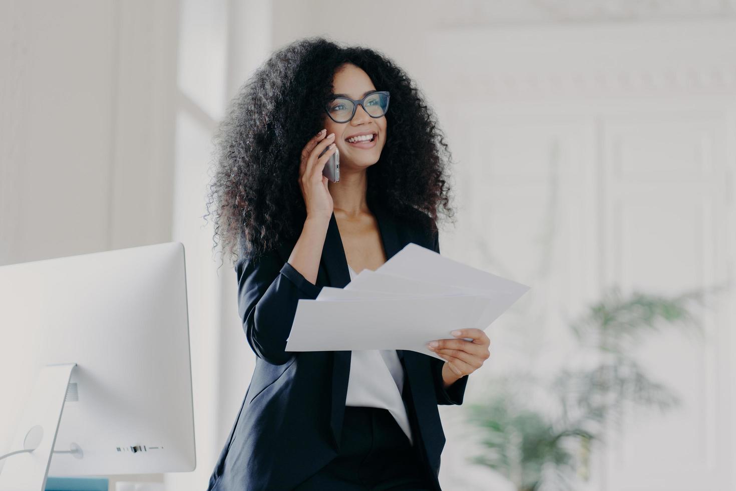 heureuse femme d'affaires prospère avec une coiffure afro porte des lunettes pour protéger les yeux, détient des documents, passe un appel téléphonique à un partenaire commercial, se tient dans une armoire près du lieu de travail. photo