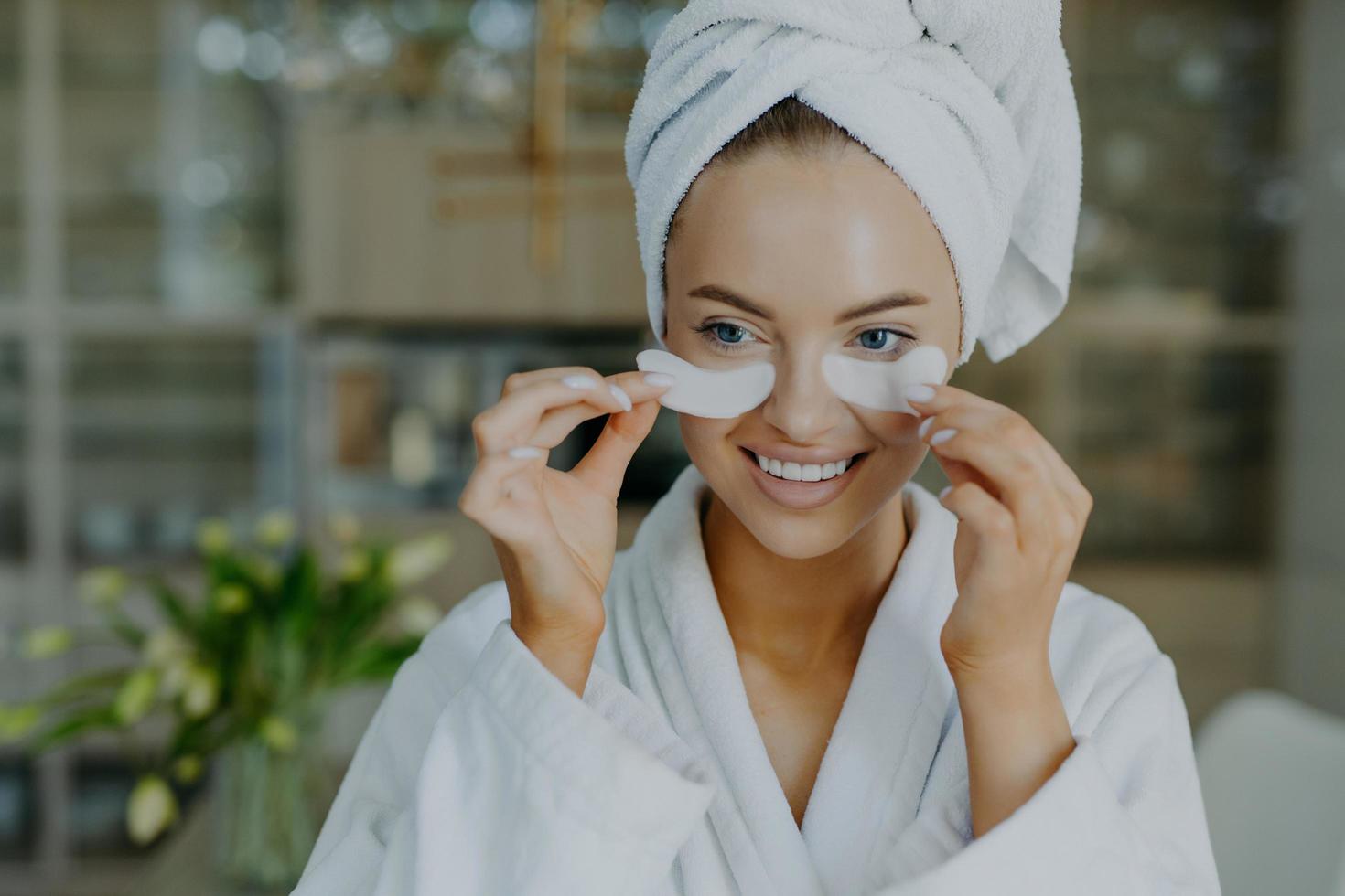 gros plan d'une belle femme européenne heureuse enlève des patchs de beauté sourit doucement a rafraîchi le teint bien soigné porte un peignoir et une serviette enveloppée subit des procédures cosmétiques à la maison photo