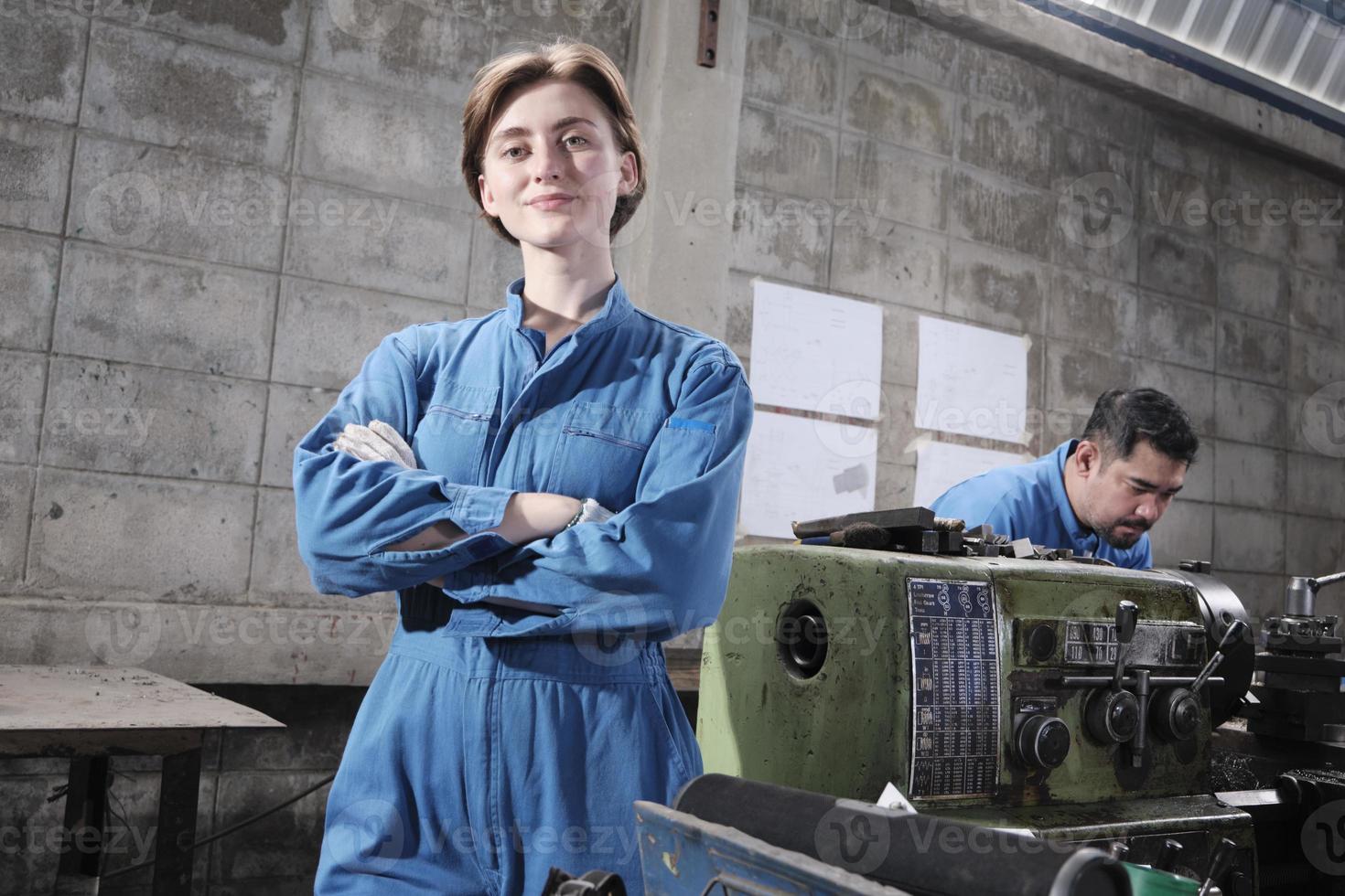 portrait d'une jeune ouvrière blanche de l'industrie se tient debout, les bras croisés, regarde la caméra et sourit, un partenaire masculin travaille avec une machine derrière elle dans une usine de fabrication, des ingénieurs mécaniques professionnels. photo