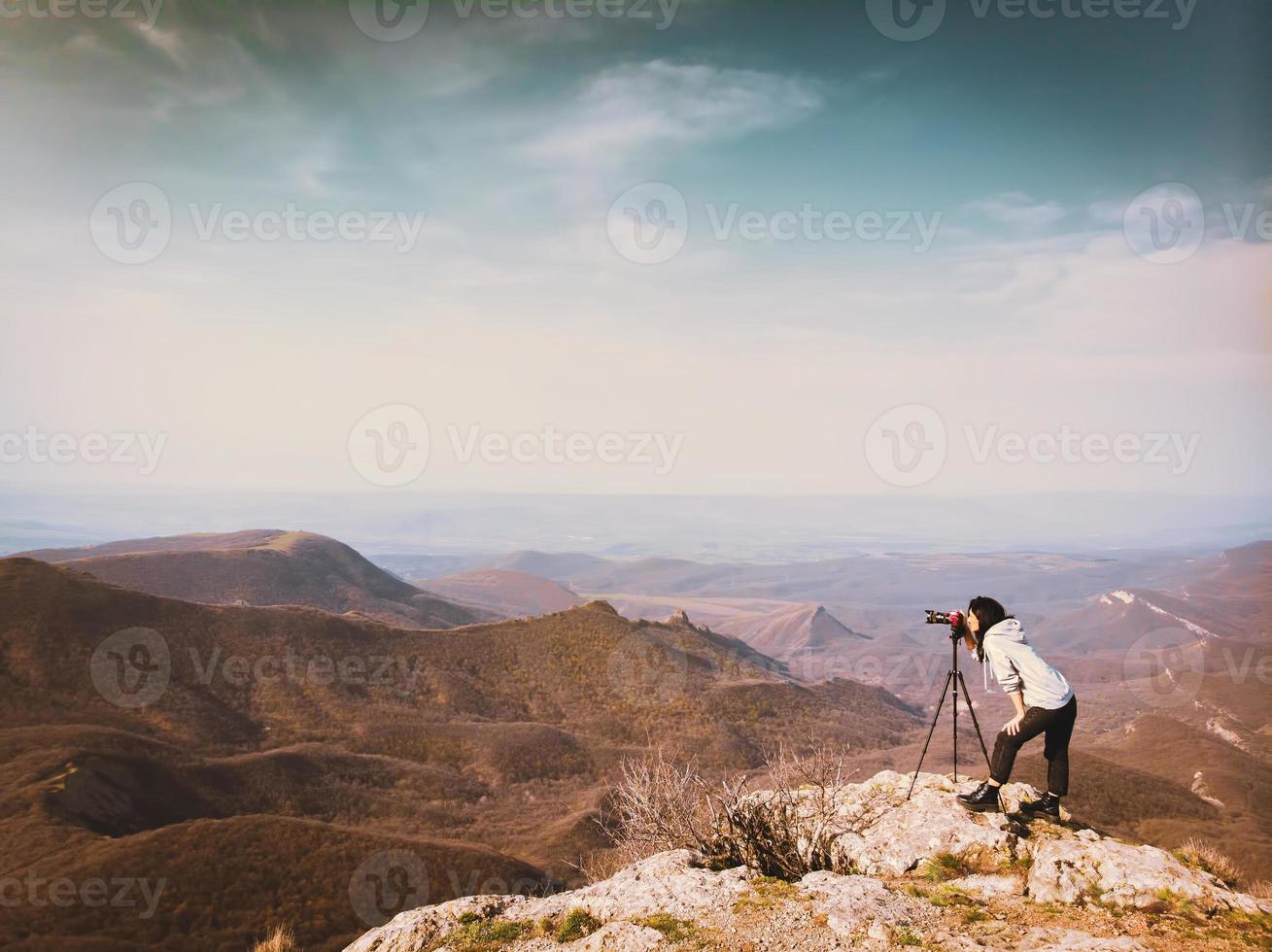 Vue latérale jeune femme hipster caucasienne attrayante créateur de contenu photographier les montagnes avec un appareil photo reflex numérique girly rouge sur trépied