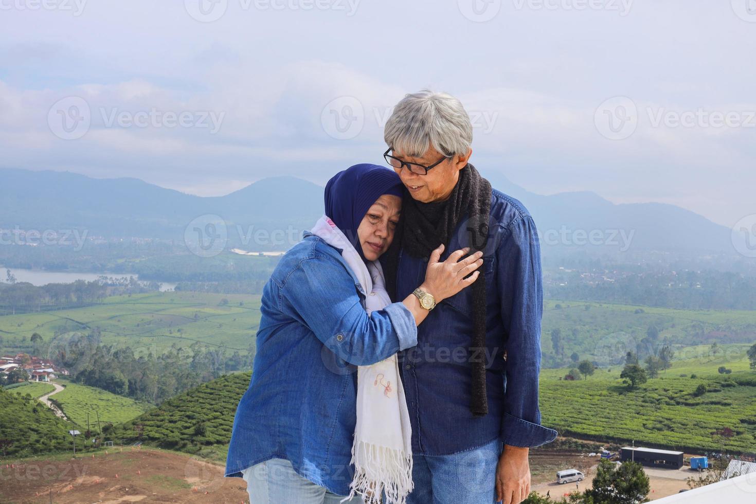 un couple de personnes âgées asiatiques dans un style décontracté est debout, une vieille femme embrasse son mari contre un paysage dramatique. vue sur la plantation de thé, la montagne et le lac. photo