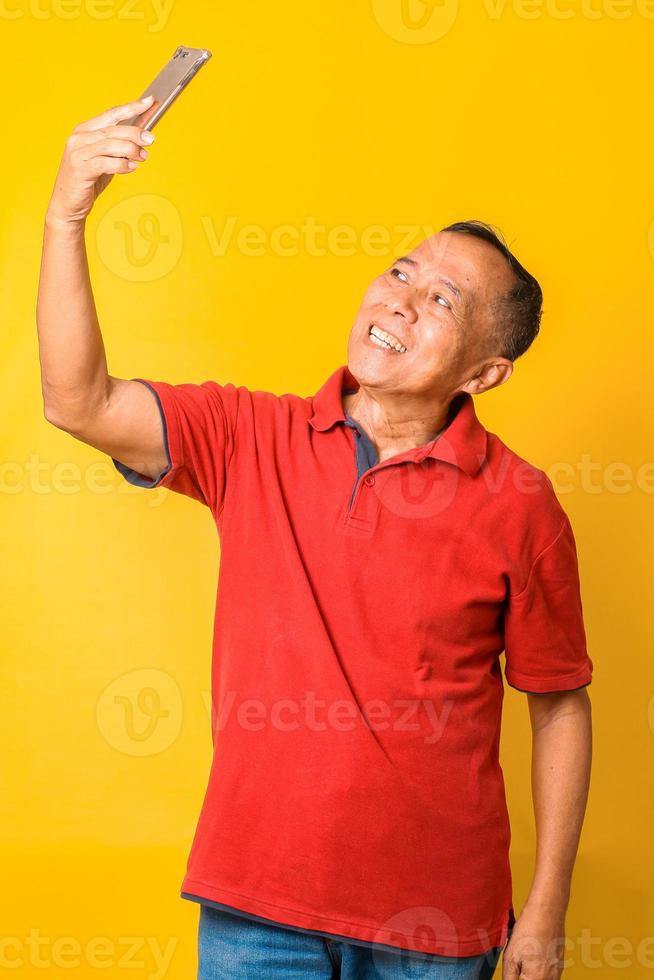 heureux homme senior asiatique prenant selfie utiliser smartphone. mature à la retraite des années 60 profitant d'un appel vidéo de réunion virtuelle, chat, parler, regarder un smartphone sur fond jaune. photo