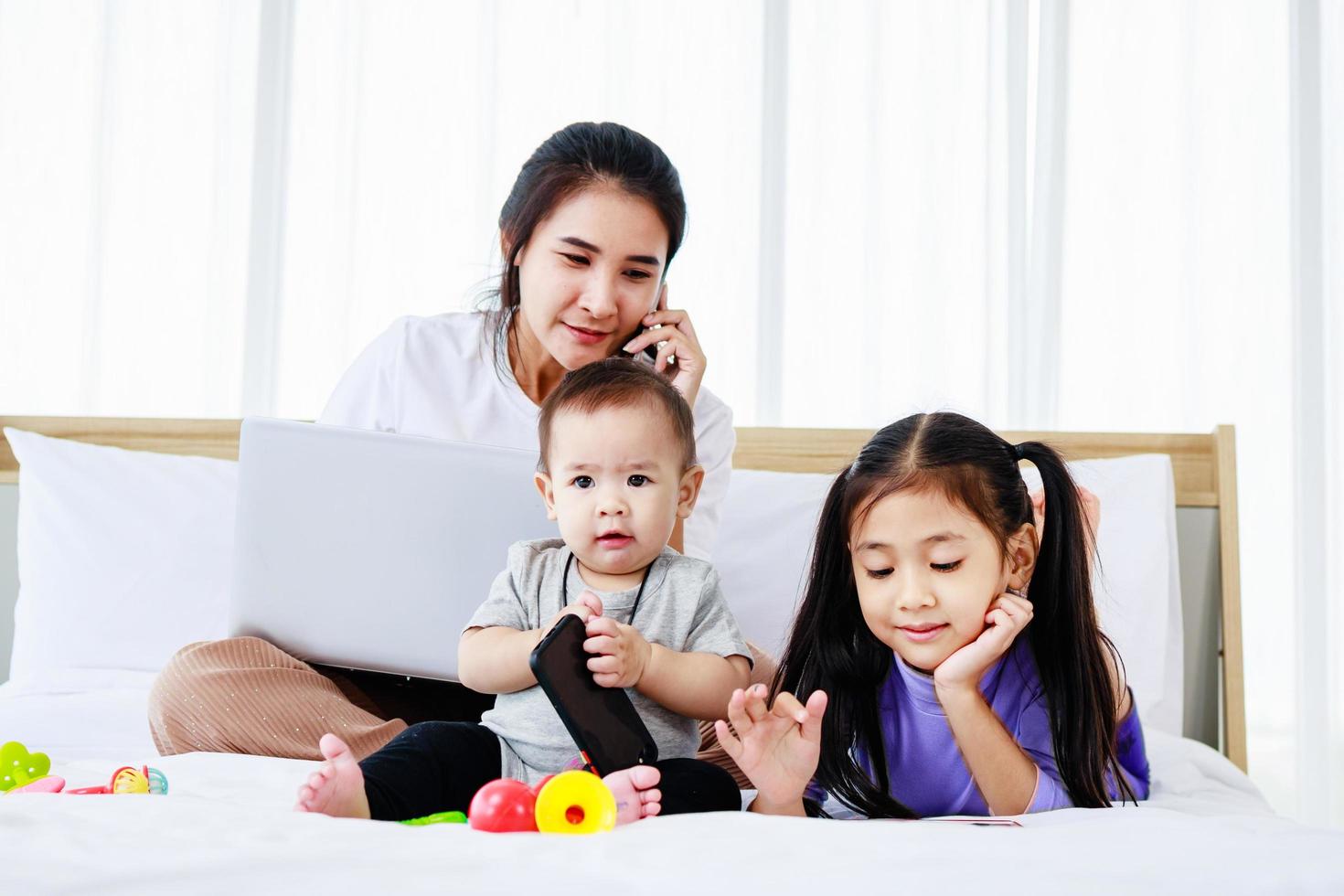 petite fille et bébé jouent ensemble aux jouets de bébé sur le lit pendant que la mère occupée travaille sur un ordinateur portable photo