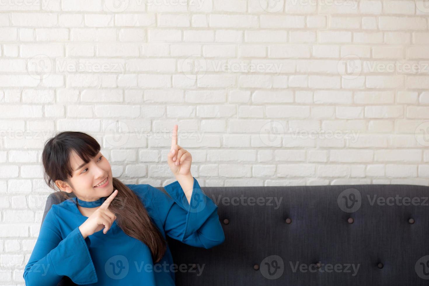 beau portrait jeune femme asiatique pointant avec espace de copie sur fond de béton de ciment, fille souriante assise sur un canapé et montrant l'avenir ou la planification avec un message de vous. photo