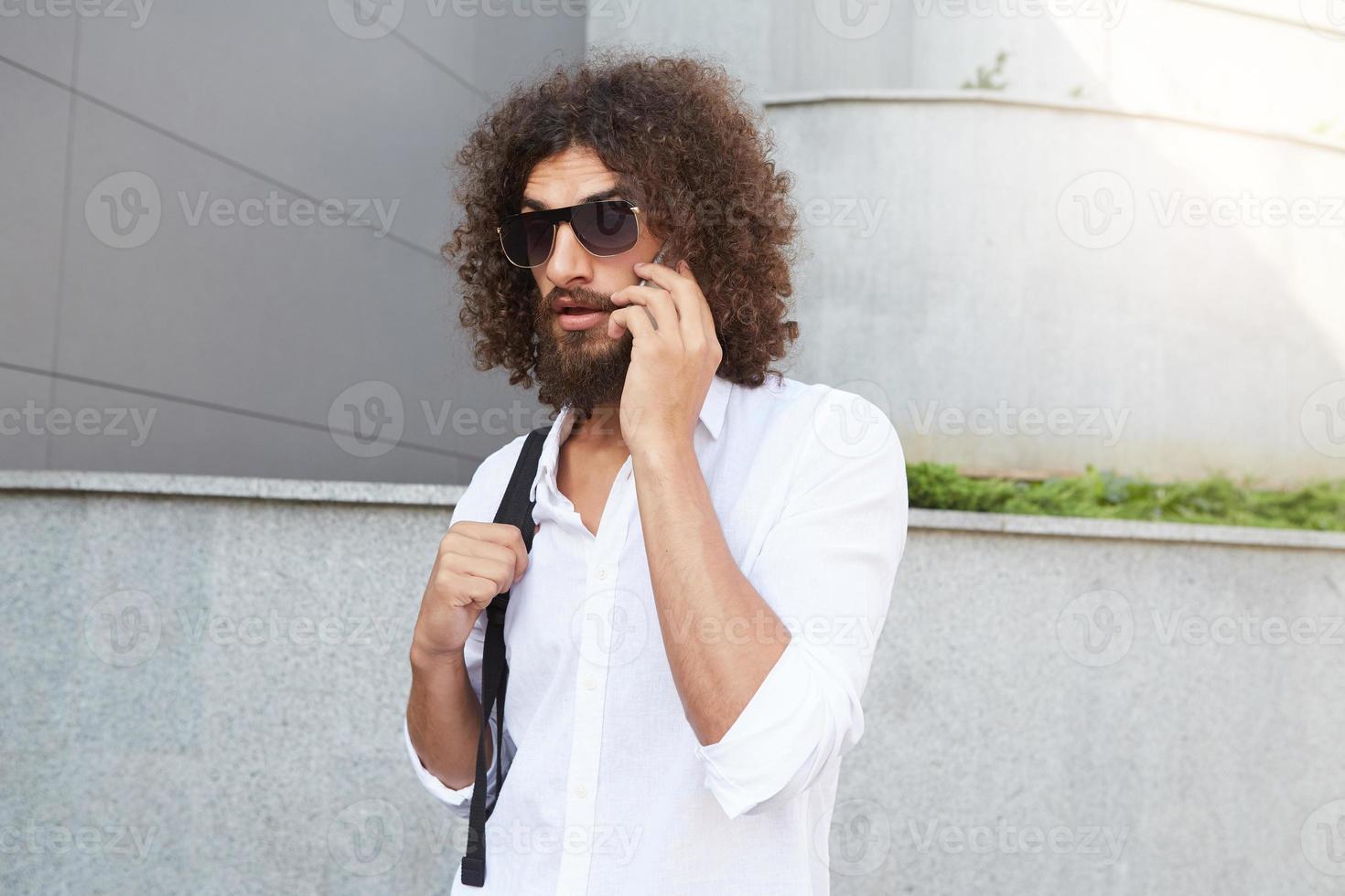 séduisant jeune homme barbu aux cheveux bouclés foncés marchant dans la rue tout en ayant une conversation téléphonique importante, portant une chemise blanche et un sac à dos noir photo