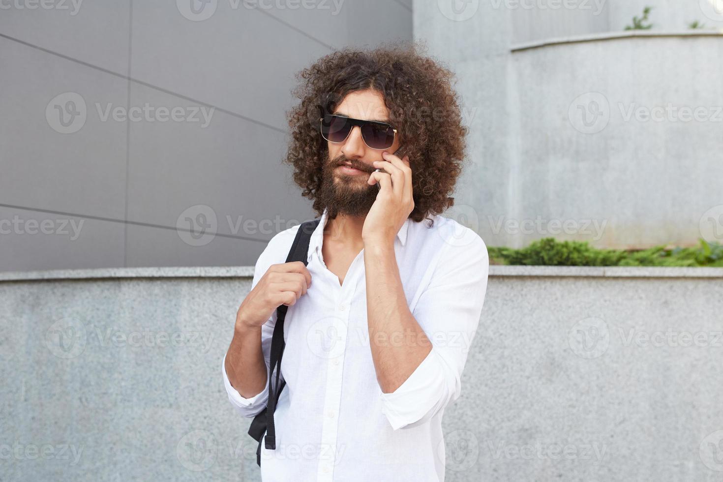 joli jeune homme bouclé avec barbe marchant dans la rue par une journée ensoleillée tout en parlant au téléphone, portant une chemise blanche et un sac à dos noir photo