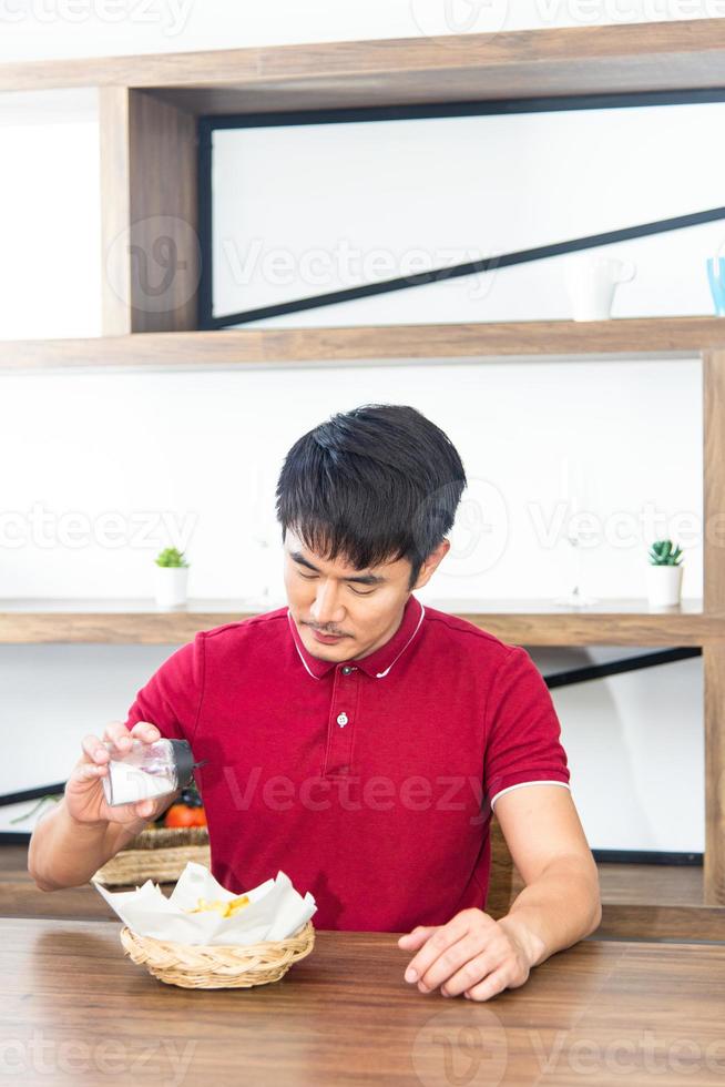 jeune homme asiatique souriant avec un t-shirt rouge décontracté aime prendre son petit déjeuner, manger des frites, jeune homme cuisiner dans la salle de cuisine de style loft photo