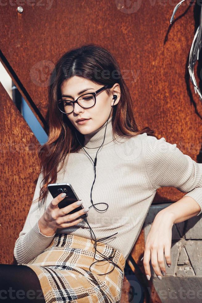 image de belle femme élégante tenant un téléphone portable. jeune fille européenne debout dans la rue et utilisant un téléphone portable. femme écoutant de la musique avec le téléphone et s'amusant. notion d'internet mobile. photo