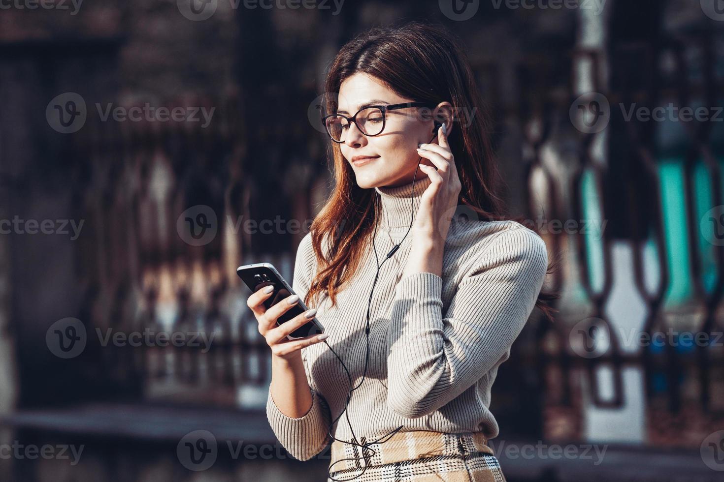 image de belle femme élégante tenant un téléphone portable. jeune fille européenne debout dans la rue et utilisant un téléphone portable. femme écoutant de la musique avec le téléphone et s'amusant. notion d'internet mobile. photo