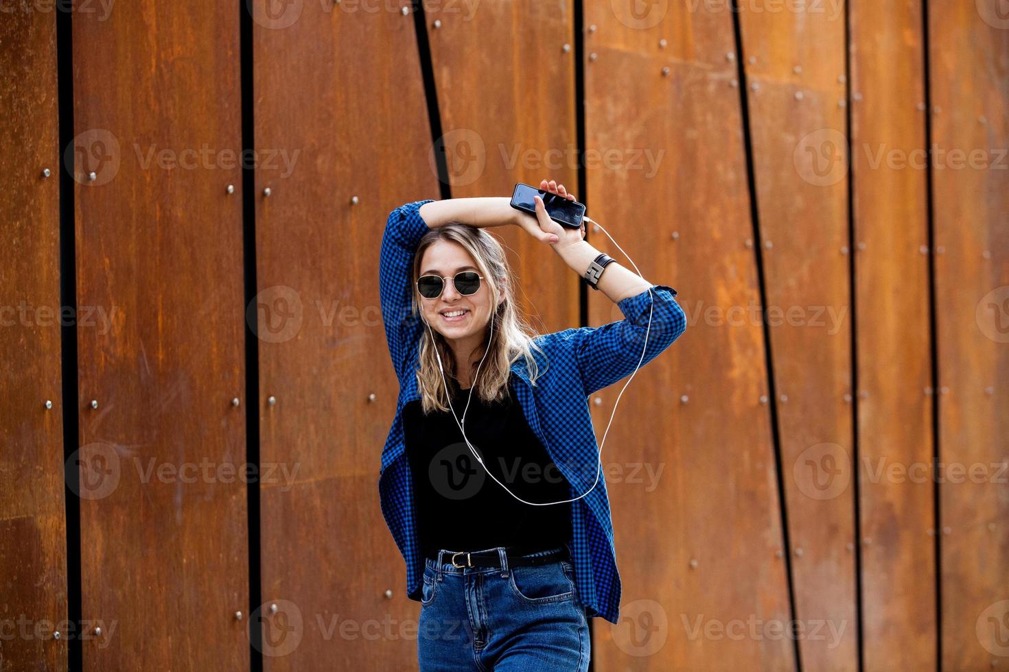 photo d'une jolie jeune étudiante souriante et joyeuse portant des lunettes de soleil à l'extérieur à l'aide d'un téléphone portable bavardant en écoutant de la musique avec des écouteurs.