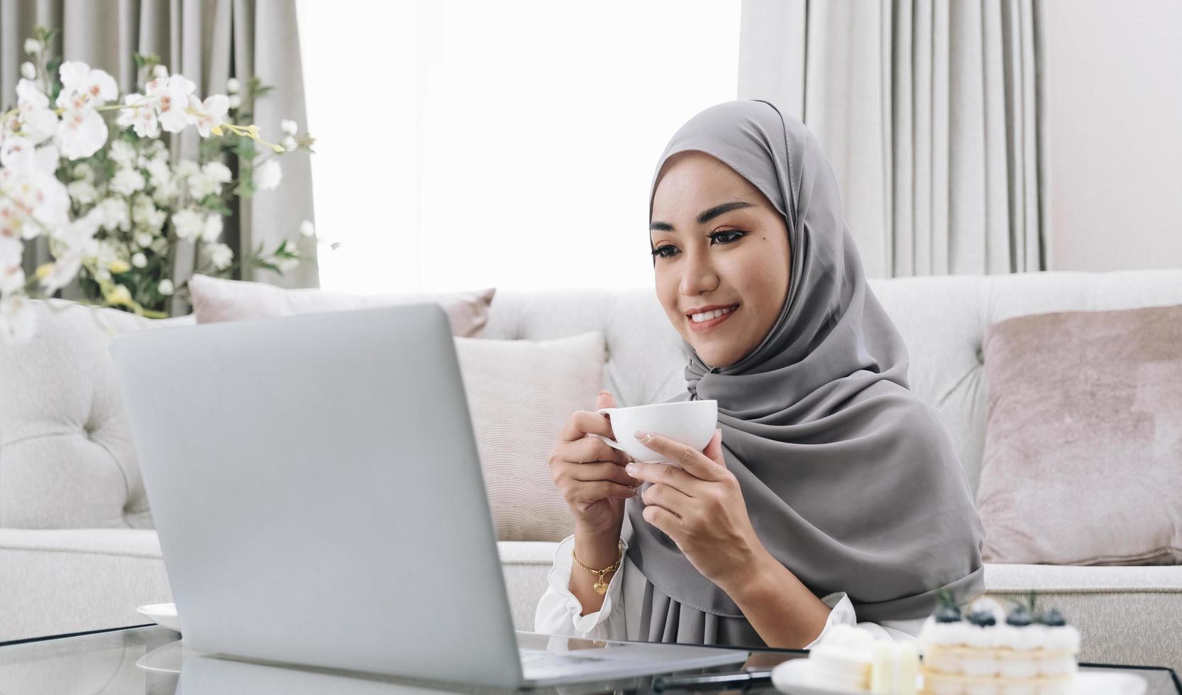 mode de vie domestique. femme musulmane heureuse utilisant un ordinateur portable à la maison, regardant un film et buvant du café, se reposant sur un canapé dans le salon, vue latérale photo