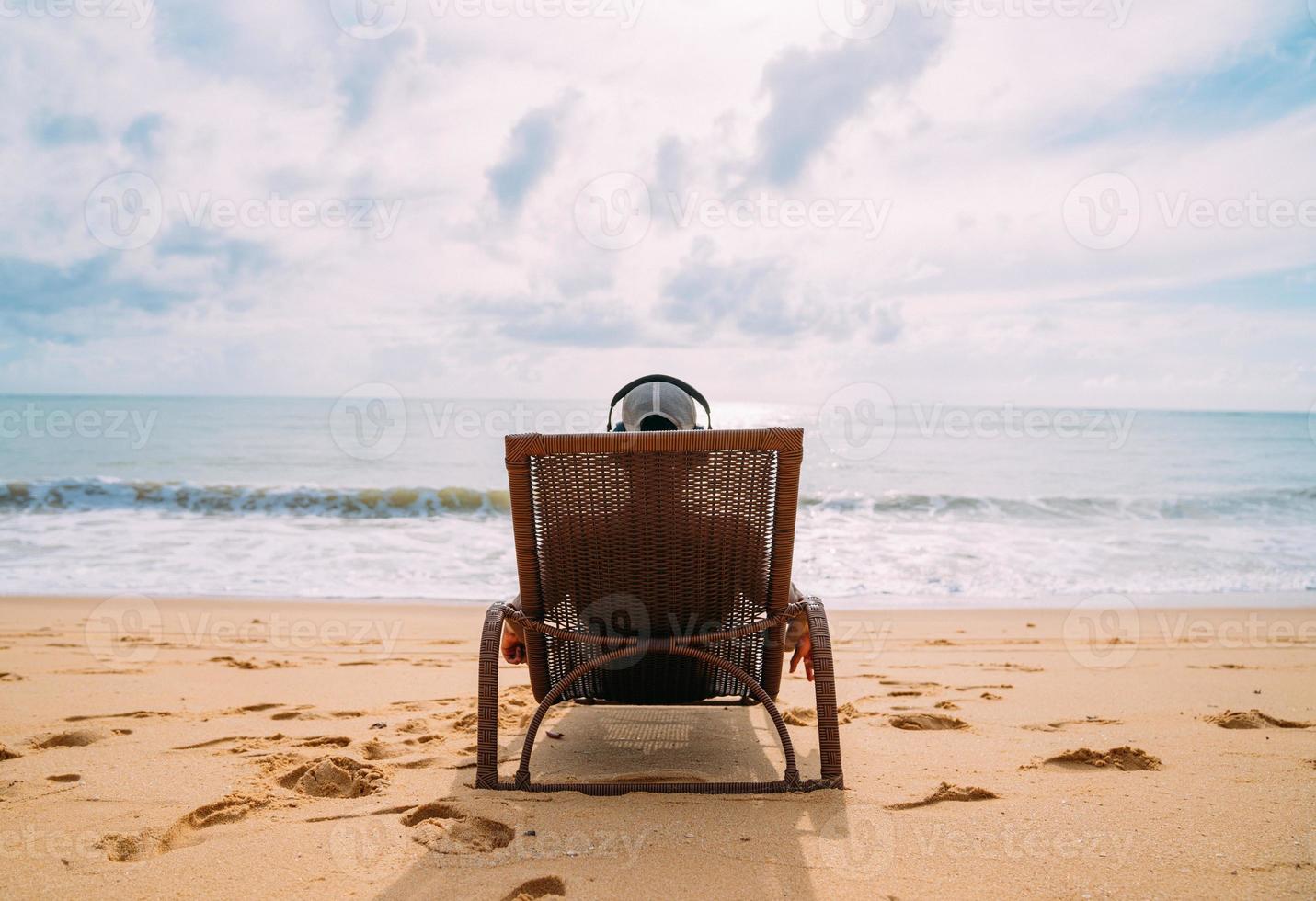 vacances d'été, technologie et concept internet. homme latino-américain assis sur la chaise de plage, écouter de la musique avec un casque et prendre le soleil sur la plage photo