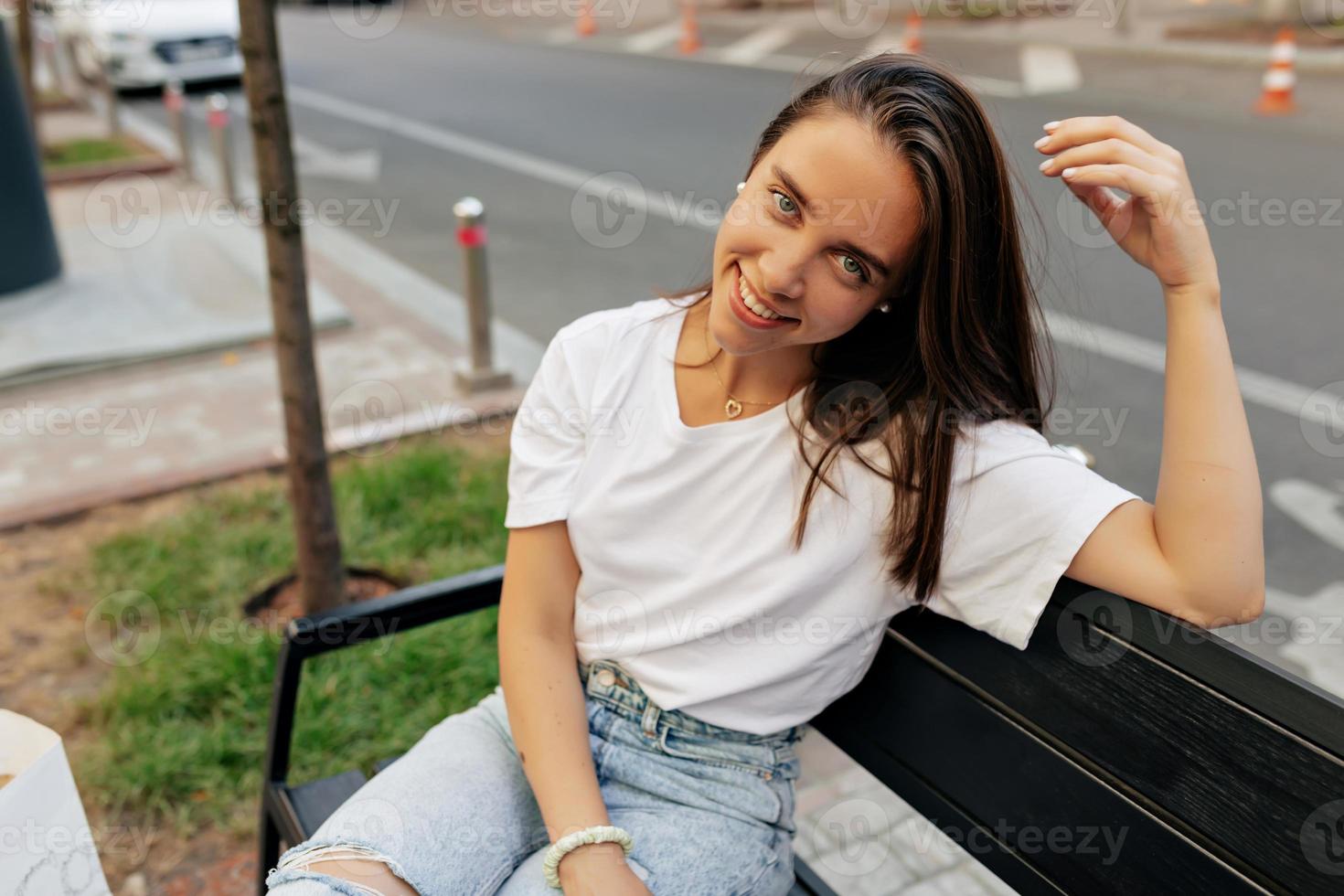 adorable jolie fille aux yeux bleus et aux cheveux noirs lâches portant un t-shirt blanc et un jean se reposant dehors sur un banc et jouant avec elle sa photo