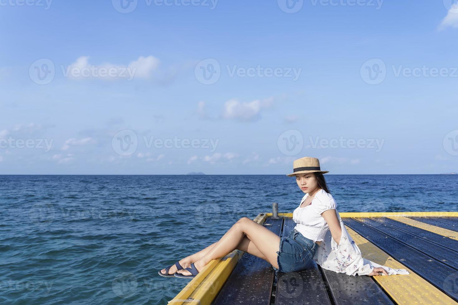 femme inquiète assise sur un pont avec un chapeau de paille océan tropical au bord de la mer dans une journée ensoleillée. fille solitaire assise seule au bord de la mer, se relaxant et pensant. photographie de concept d'émotion humaine photo