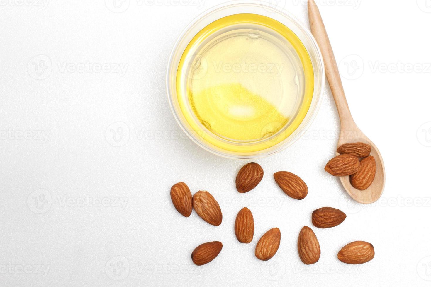 huile d'amande jaune claire dans un bol transparent avec une cuillère en bois dessus et plusieurs amandes sur fond blanc.vue de dessus, à plat, de haut en bas. photo