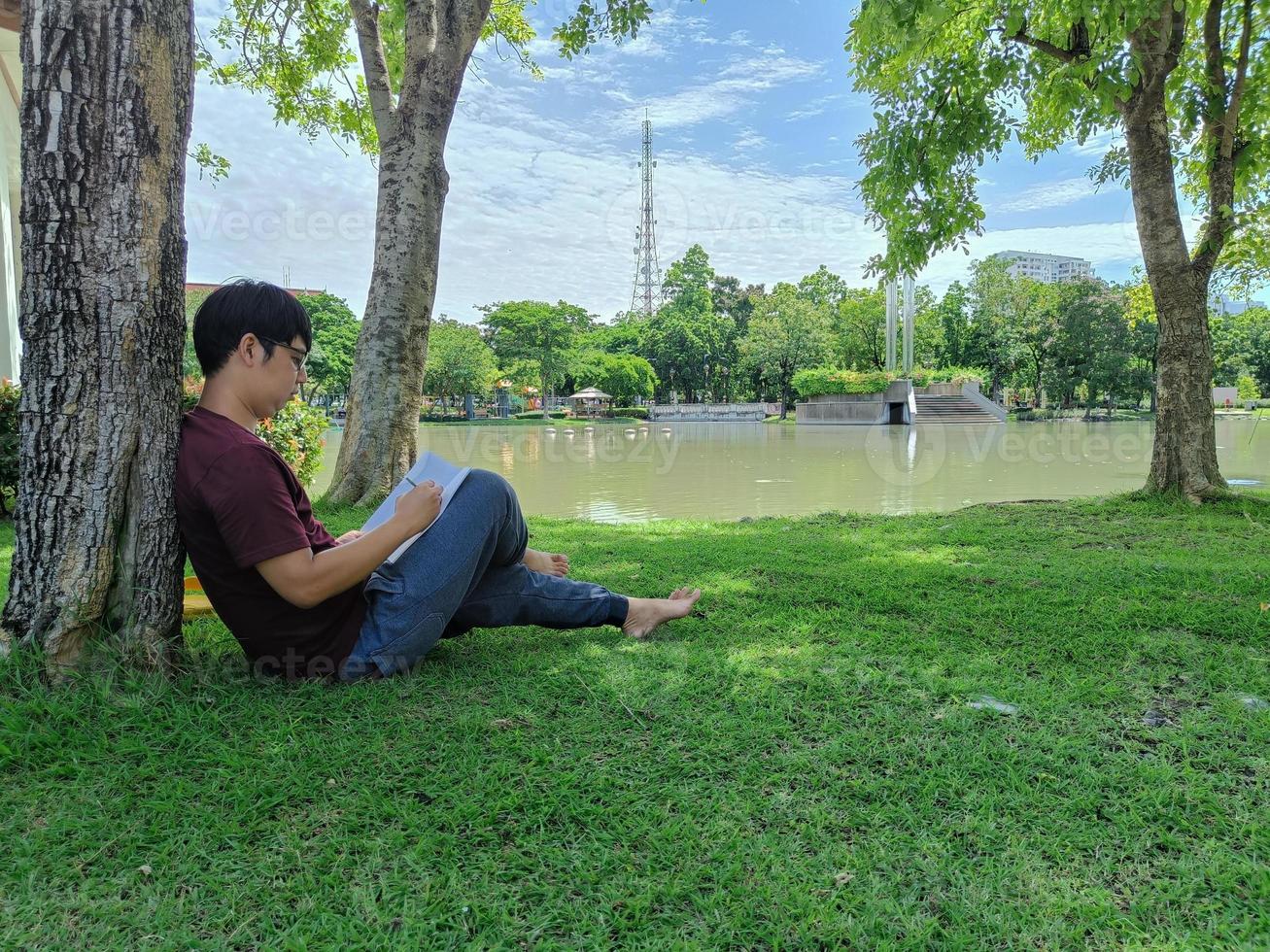 jeune homme asiatique portant des lunettes aux cheveux noirs est assis sous un arbre tenant un livre. heureux et se détendre. à quoi pense-t-on dans la ville du parc d'été photo