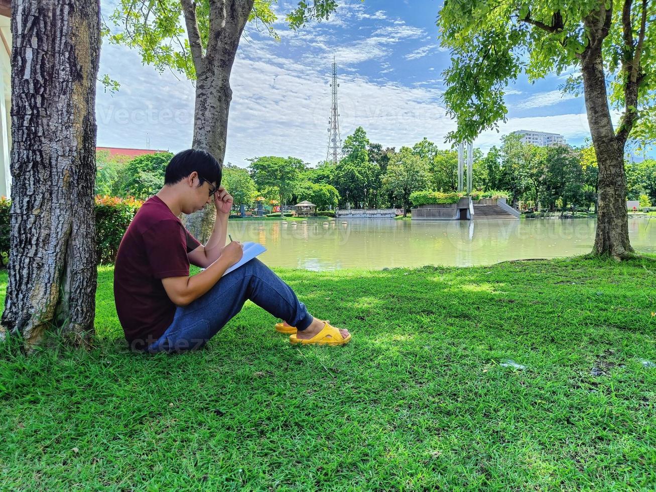 jeune homme asiatique portant des lunettes aux cheveux noirs est assis sous un arbre tenant un livre. stress et anxiété. à quoi tu penses dans la ville du parc d'été photo