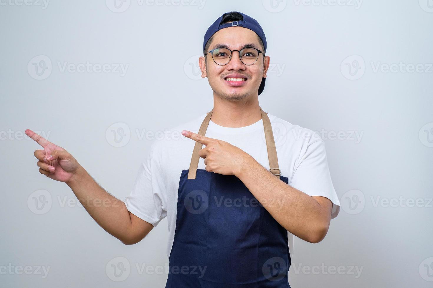 jeune et beau barista asiatique portant un tablier pointant quelque chose sur son côté avec ses deux mains photo