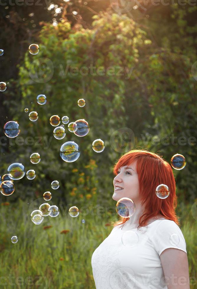 jeune femme en t-shirt blanc joue avec des bulles de savon. heureuse fille rousse au soleil. photo