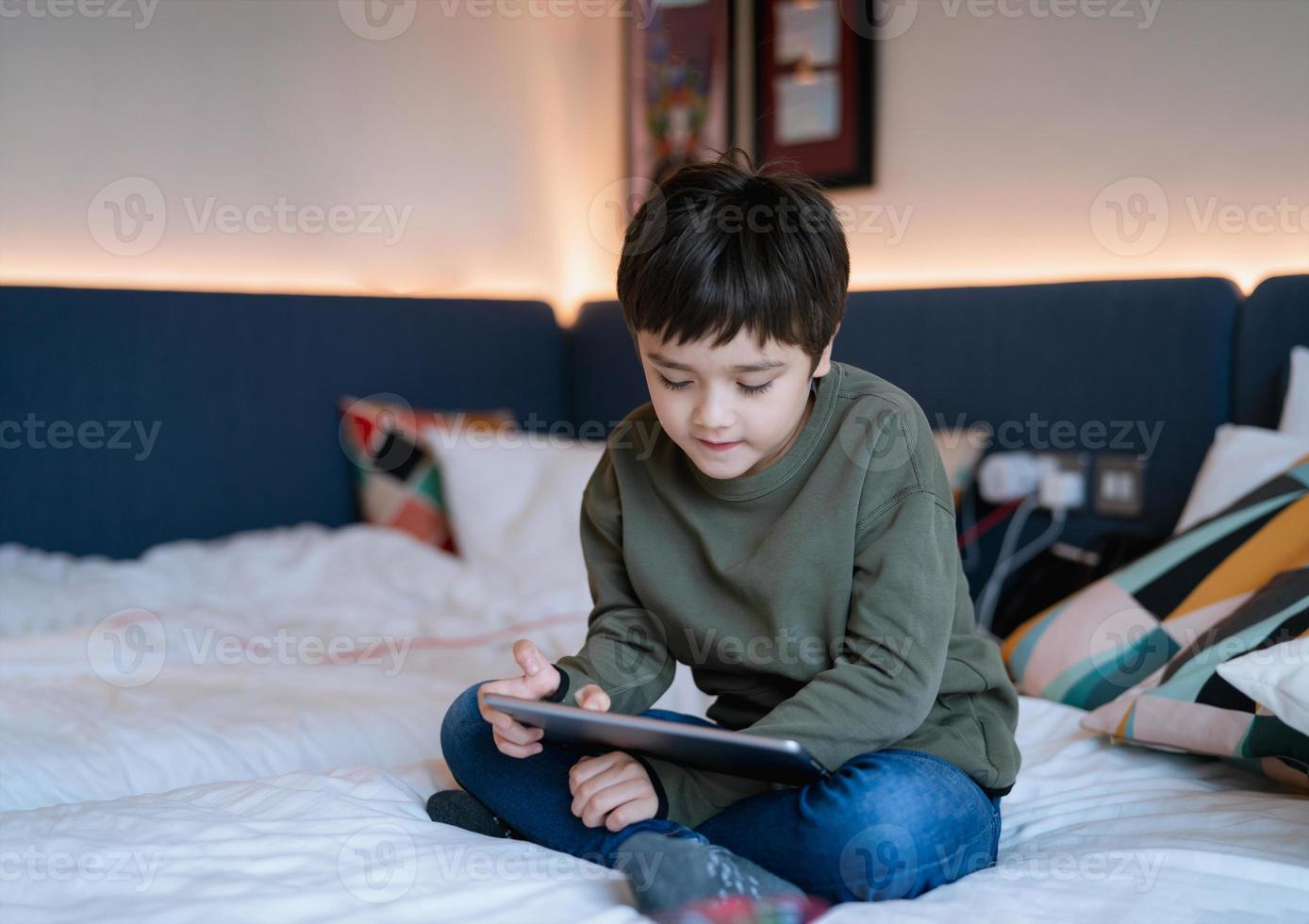 enfant regardant un dessin animé et discutant avec des amis sur une tablette, garçon heureux assis dans son lit jouant à des jeux en ligne sur un pavé numérique, portrait enfant se relaxant à la maison dans sa chambre le week-end. photo