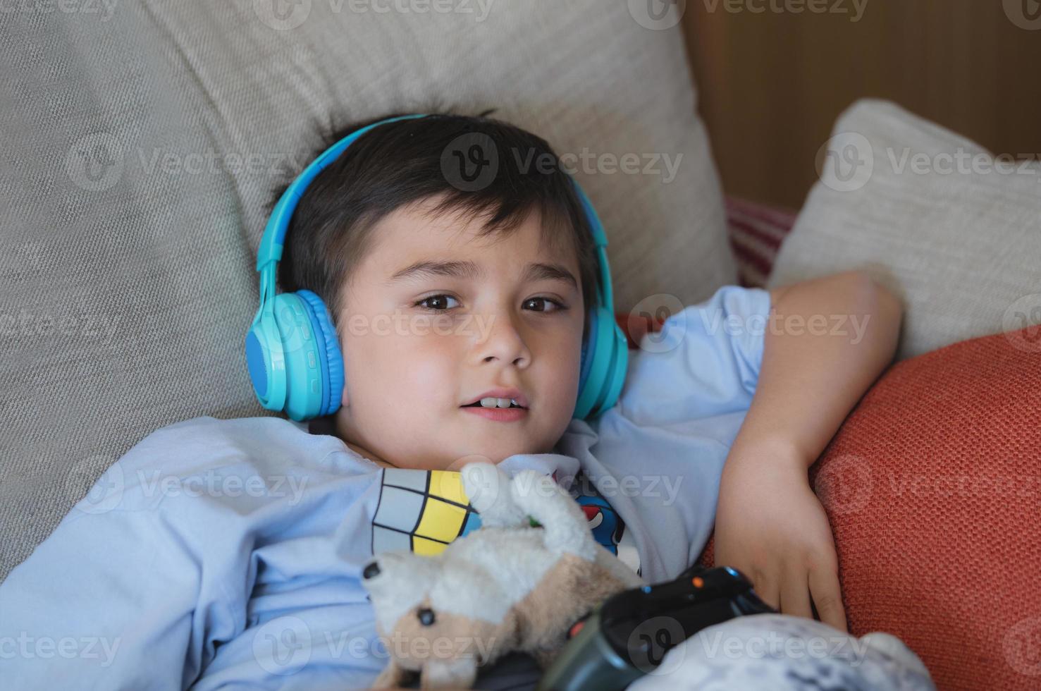 portrait d'enfant portant des écouteurs et jouant au jeu vidéo. enfant tenant une console de jeu en ligne avec des amis à la maison, jeune garçon assis sur un canapé s'amusant et se relaxant seul le week-end photo