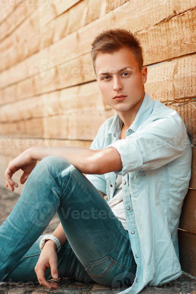 jeune bel homme en vêtements de jeans assis près d'un mur en bois. photo