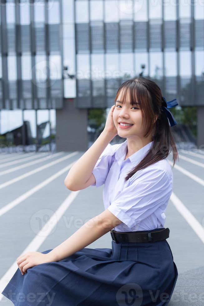 belle lycéenne asiatique en uniforme scolaire avec des sourires confiants pendant qu'elle regarde la caméra avec bonheur avec le bâtiment en arrière-plan. photo