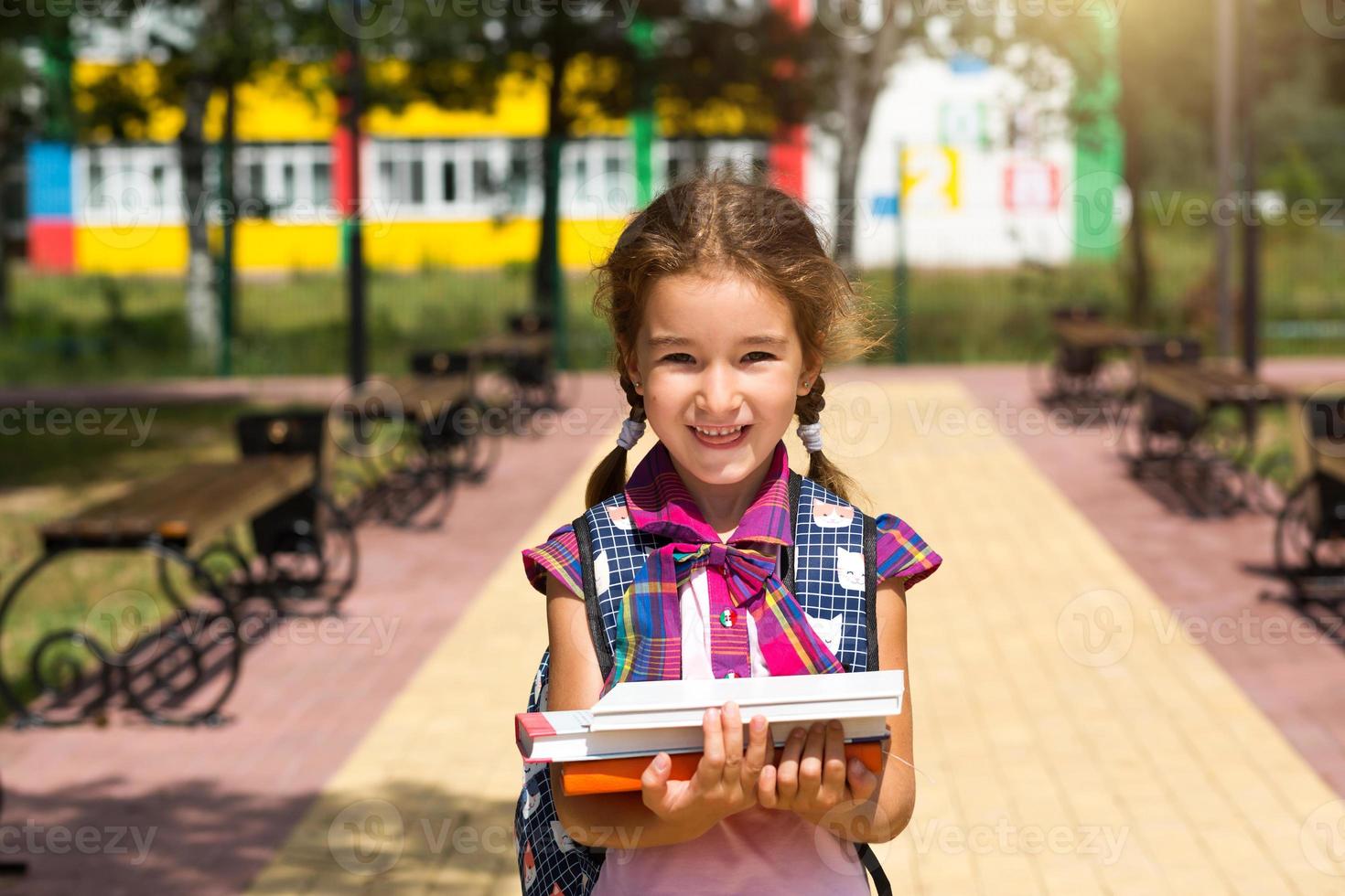 fille avec un sac à dos et une pile de livres près de l'école. retour à l'école, élève heureux, manuels lourds. éducation, classes primaires, rentrée scolaire, 1er septembre photo