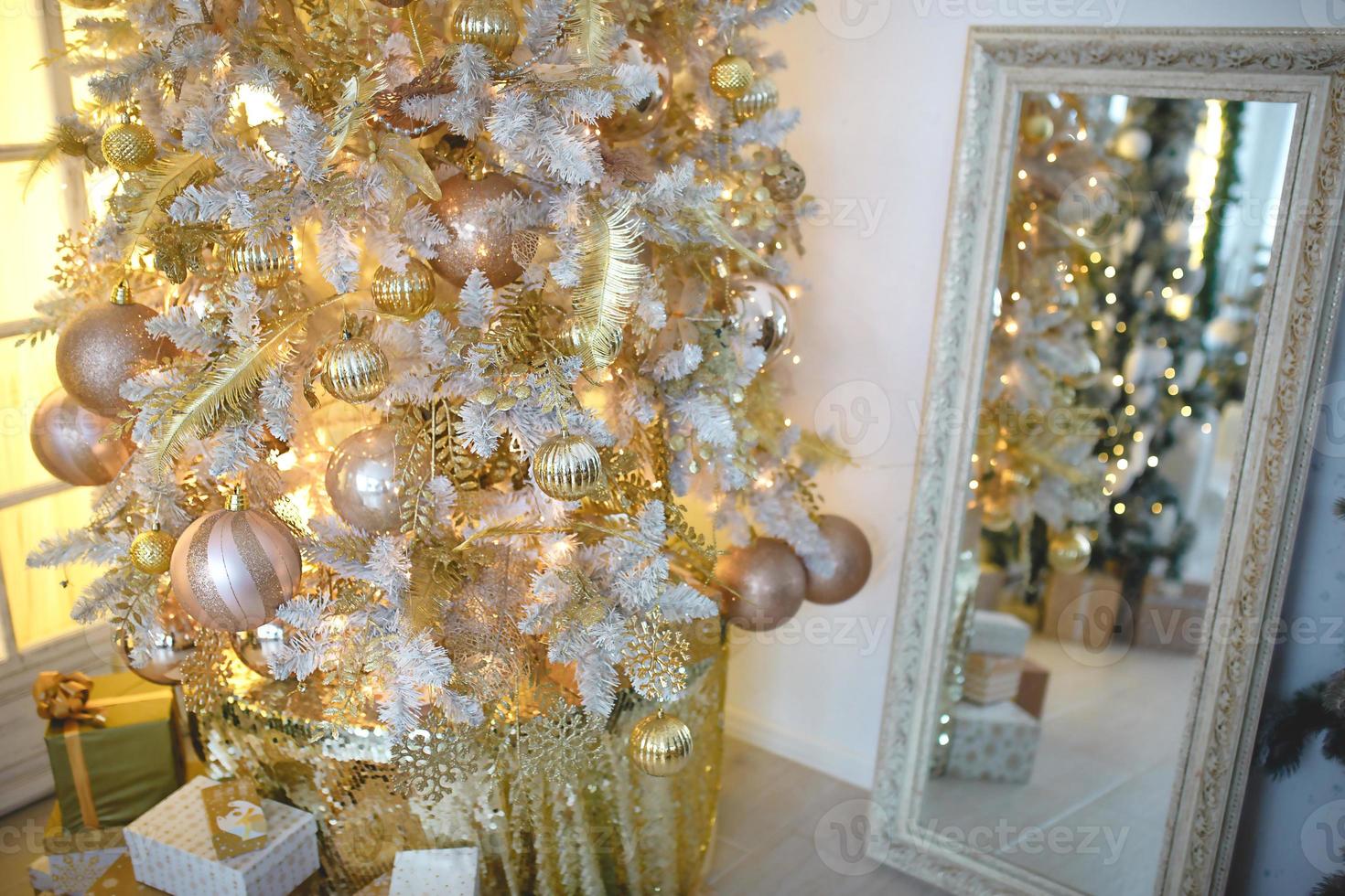 un arbre de noël avec une décoration dorée se reflète dans le miroir. nouvel an, décoration de la maison. grande fenêtre avec lumière jaune chaude, confort dans le salon photo