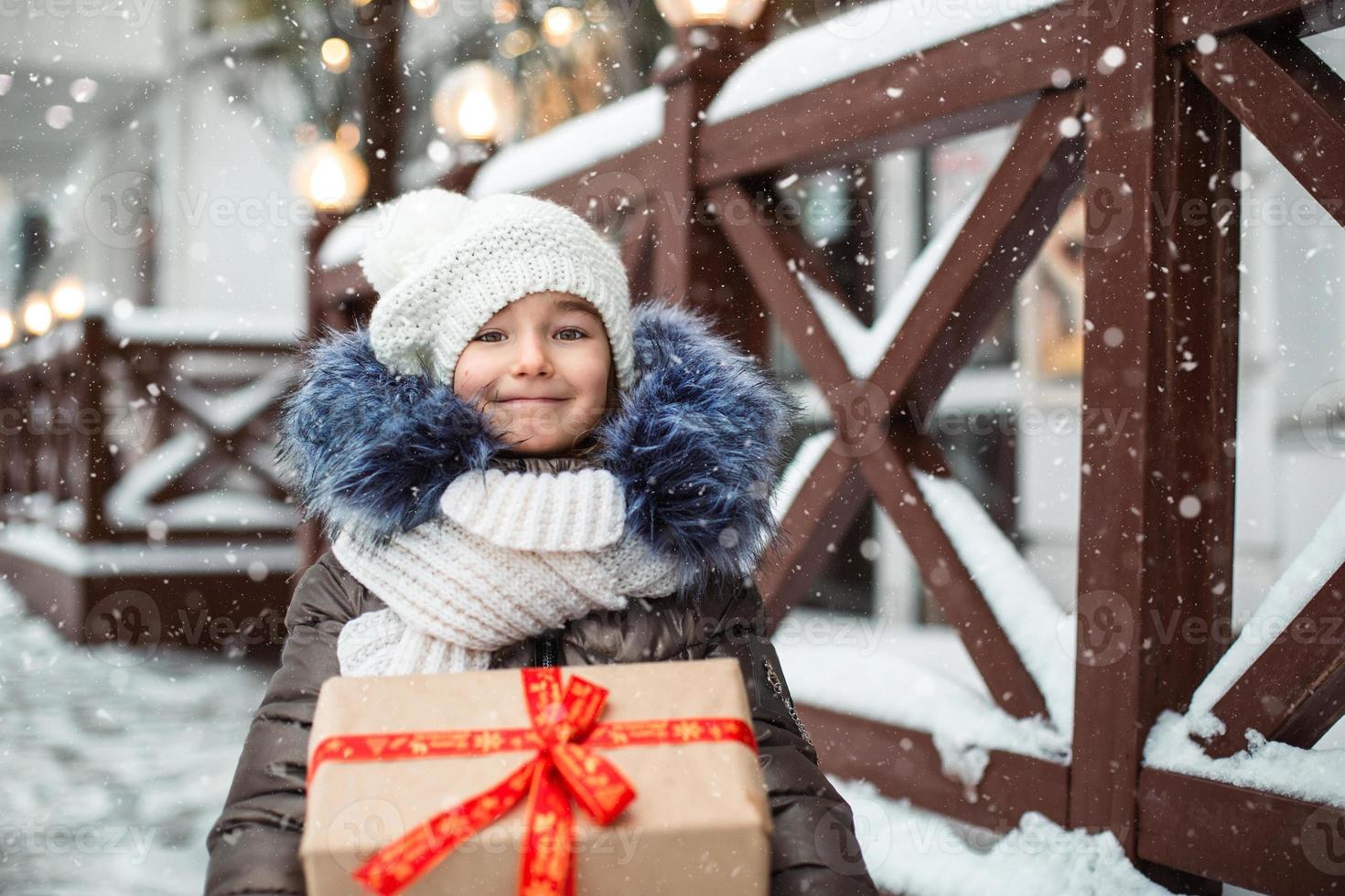 portrait d'une fille joyeuse avec une boîte-cadeau pour noël dans une rue de la ville en hiver avec de la neige sur un marché festif avec des décorations et des guirlandes lumineuses. vêtements chauds, bonnet, écharpe et fourrure. nouvel An photo