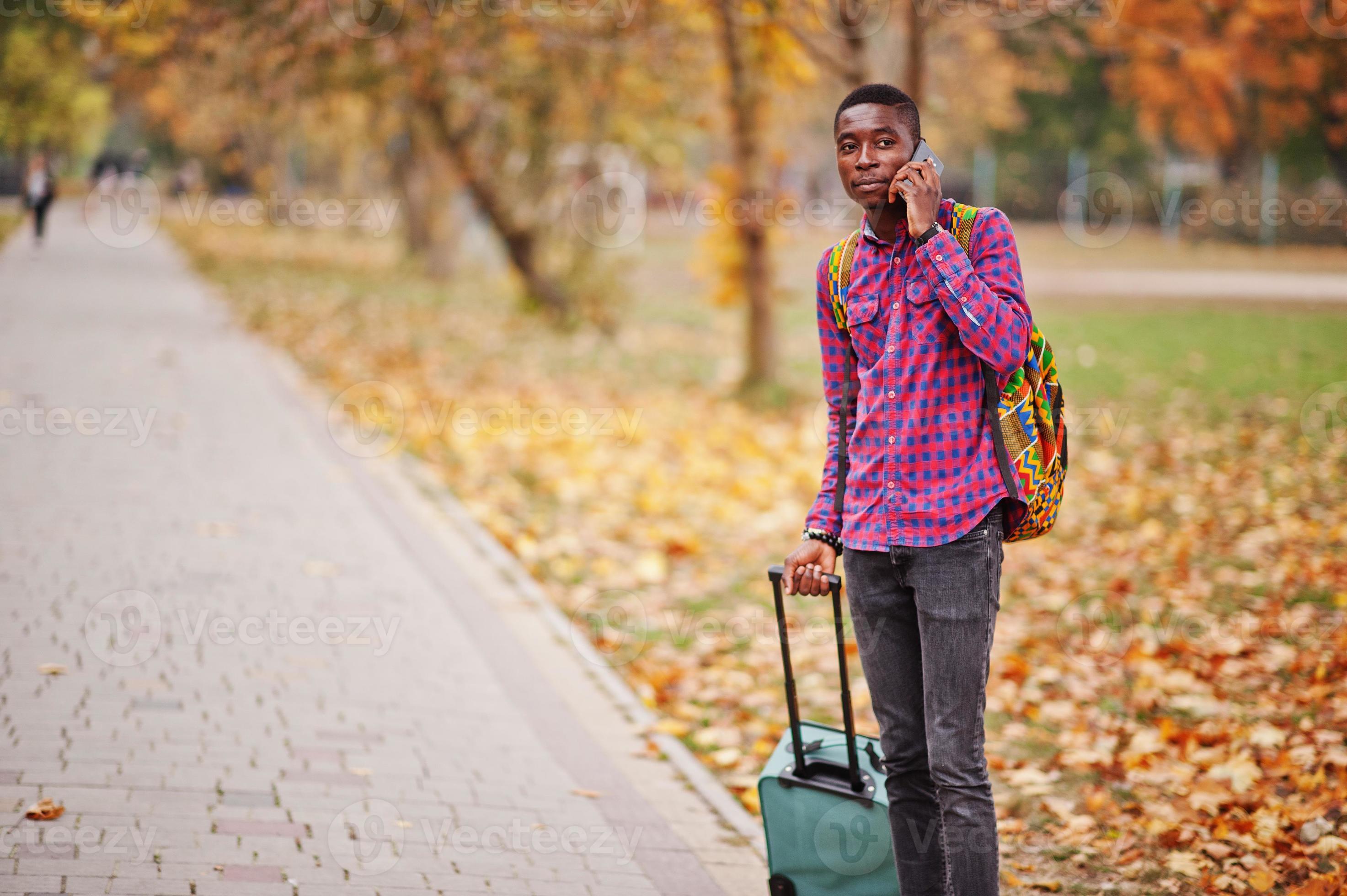 homme afro-américain en chemise à carreaux, avec valise et sac à dos.  voyageur d'homme noir au parc d'automne parlant au téléphone portable.  10570233 Photo de stock chez Vecteezy