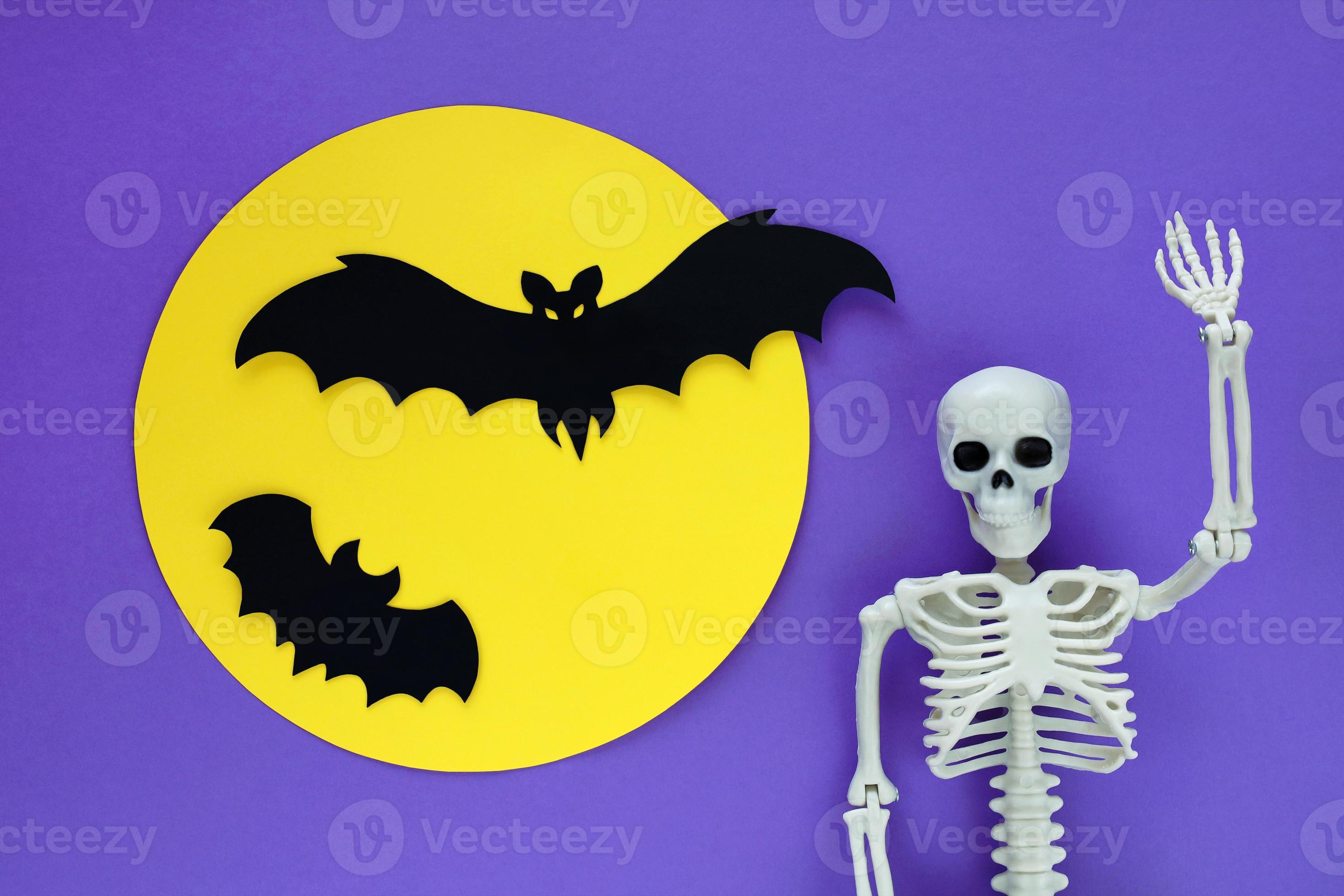 squelette sur fond d'halloween en papier violet et silhouettes de deux  chauves-souris coupées en papier noir volant sur la pleine lune jaune.  modèle anatomique en plastique squelette humain a levé la main