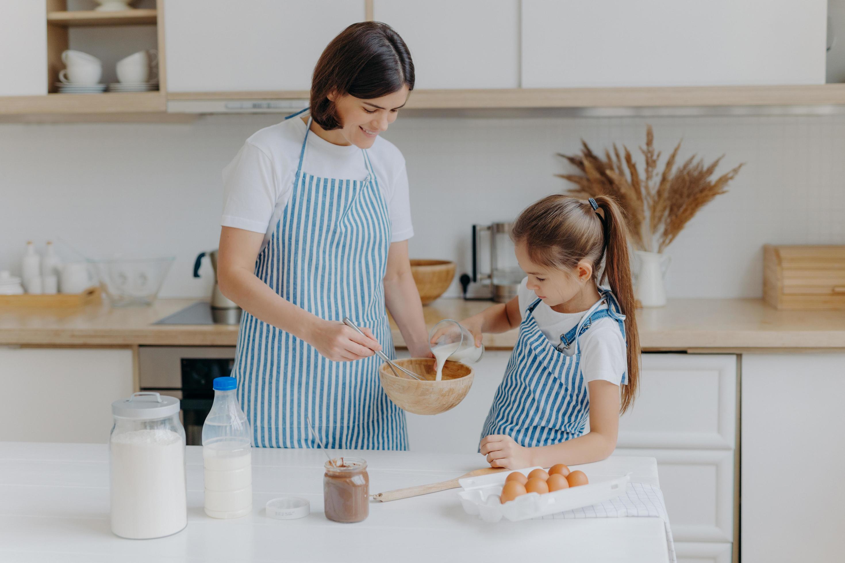 heureuse mère et fille cuisent ensemble dans la cuisine, utilisent  différents ingrédients, portent des tabliers, se tiennent contre  l'intérieur de la cuisine, la fille verse du lait à l'arc. maman  attentionnée apprend