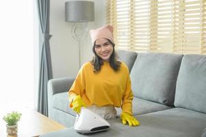 jeune femme heureuse portant des gants jaunes et passant l'aspirateur sur un canapé dans le salon.