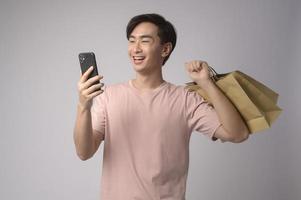 jeune homme asiatique tenant un smartphone et un sac à provisions sur fond blanc concept de studio, de shopping et de finance.